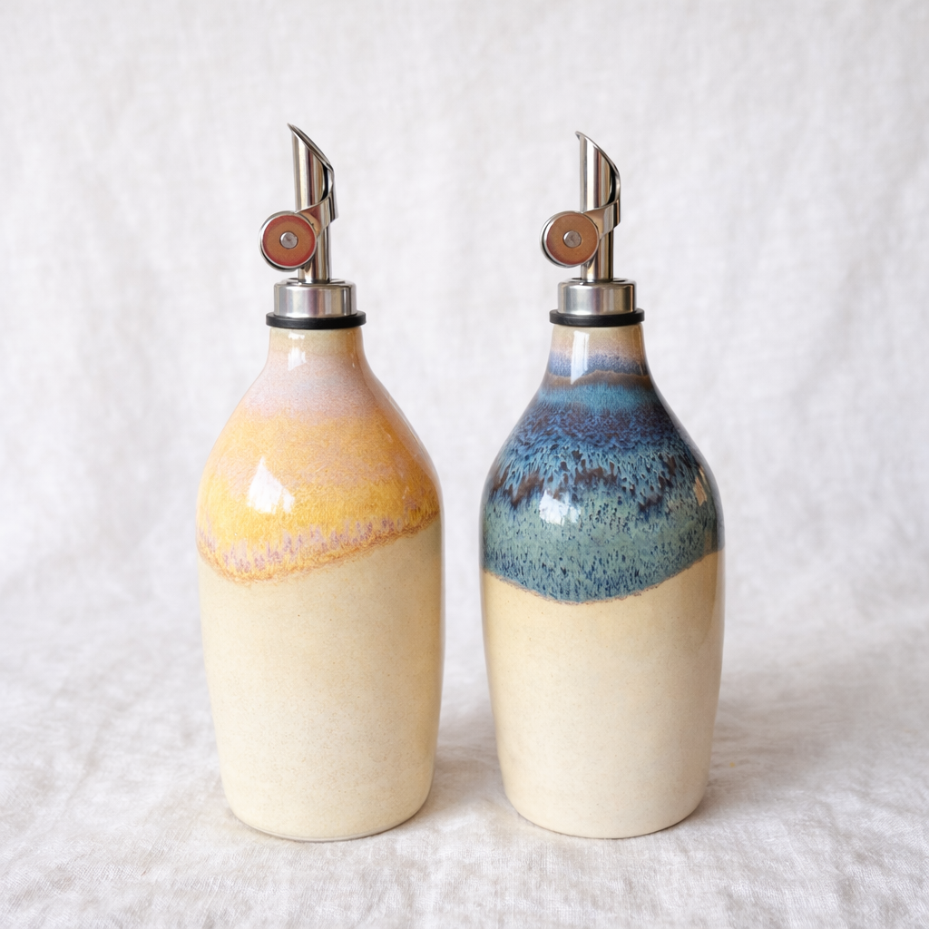 Two ceramic oil dispensers with metal spouts, one with a yellow and pink design and the other with a blue and green design, on a light fabric surface.
