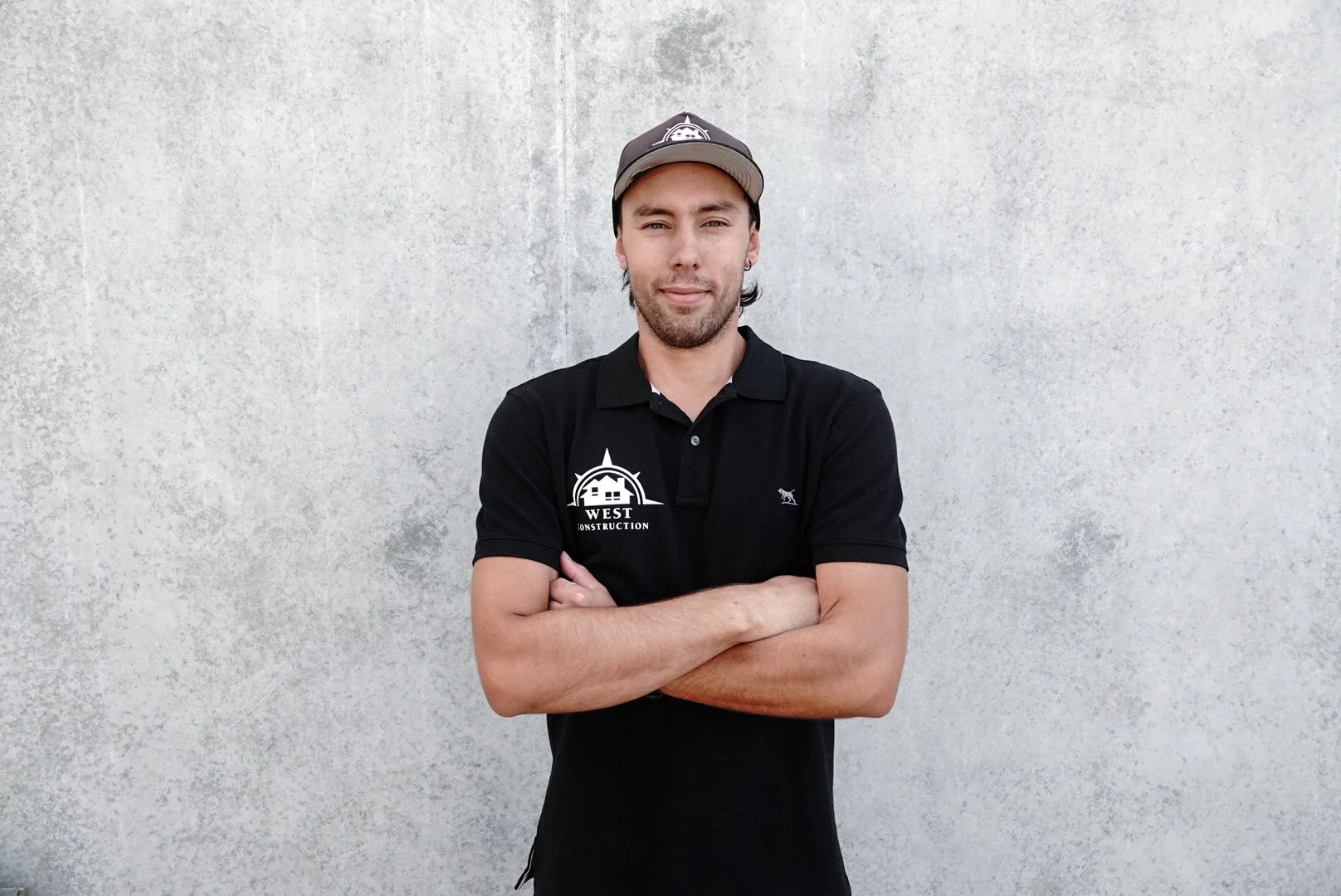 Young man with crossed arms standing against a plain concrete wall, wearing a black polo shirt with a company logo and a baseball cap.