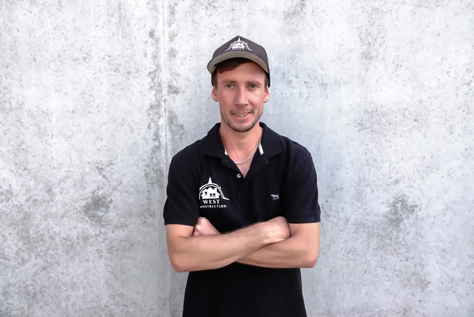Young man standing with arms crossed in front of a plain white wall, wearing a black polo shirt and a black cap, smiling slightly.