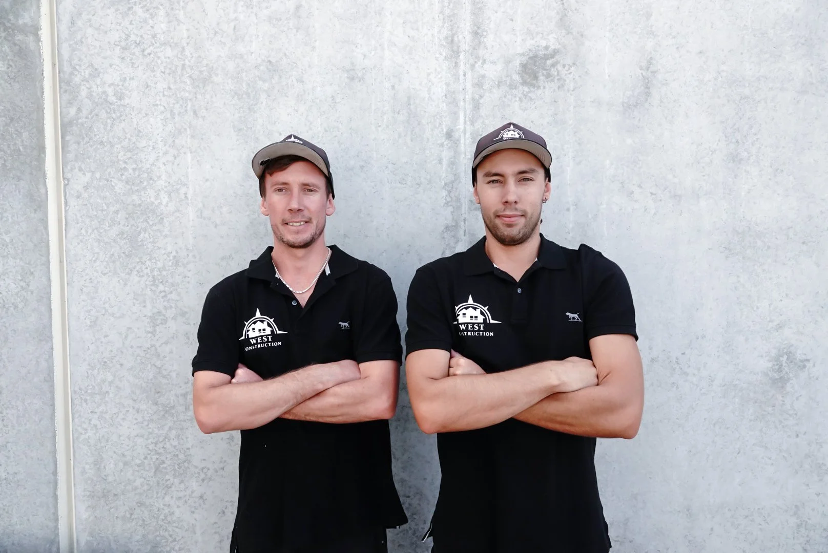 Two men standing side by side with arms crossed in front of a plain, light-colored wall. They are wearing black polo shirts with a logo and text that reads 'West Construction' and baseball caps.