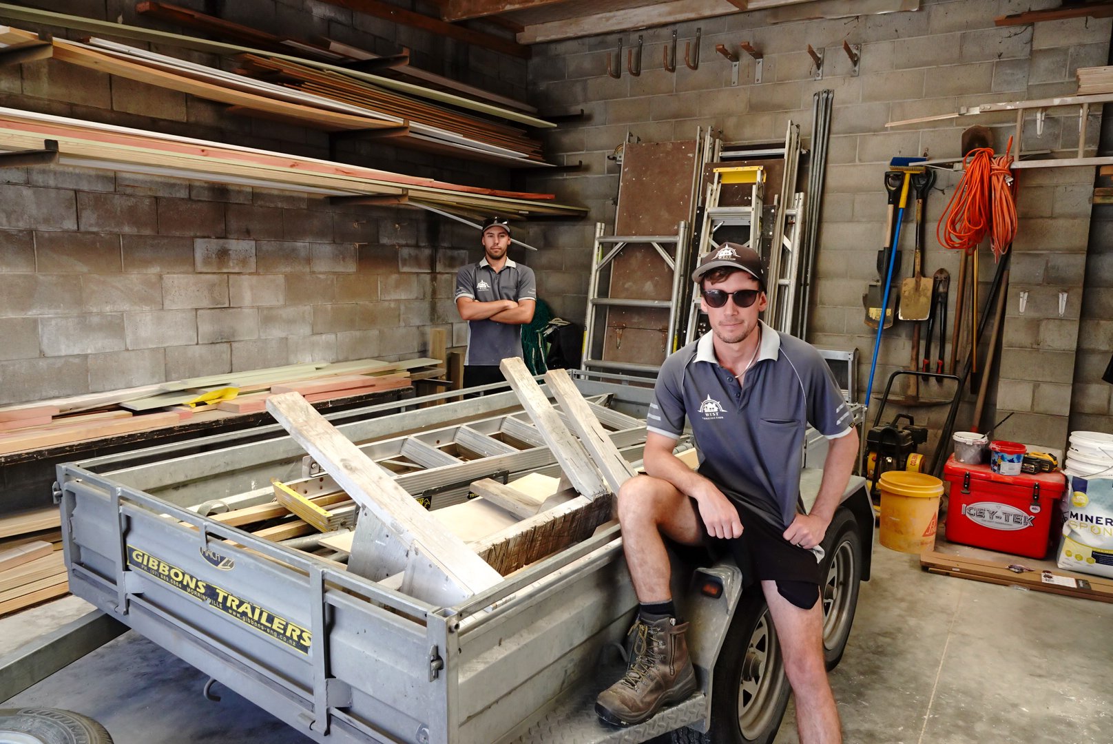 Two men in a garage or workshop, one sitting on a trailer and another standing behind, surrounded by tools and building materials.