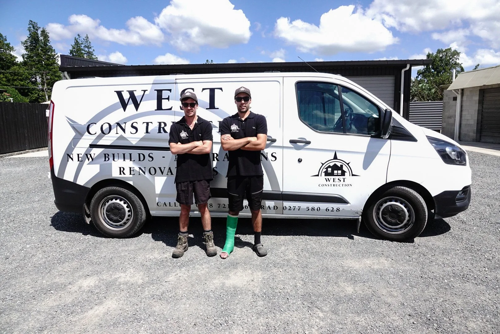 Two construction workers standing in front of a white company van with company logo and contact information, on a gravel driveway with houses and trees in the background under a partly cloudy sky.