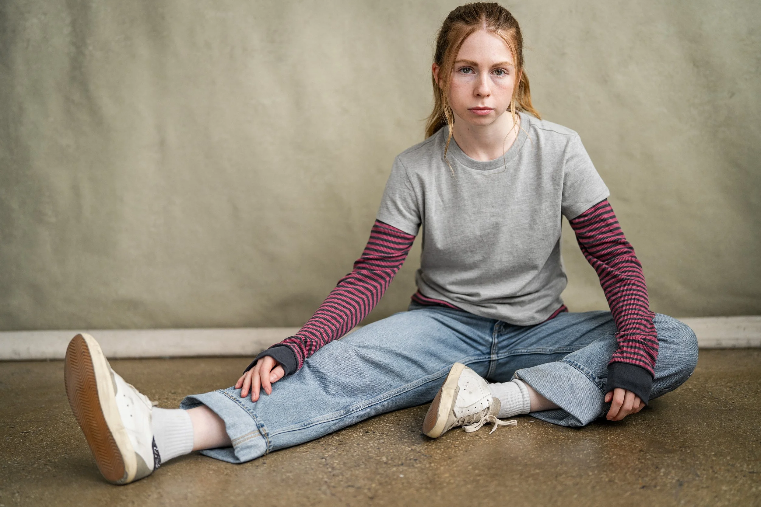 A young woman with red hair sitting on the floor with her legs stretched out, wearing a gray t-shirt with long sleeves, blue jeans, and white sneakers. Stranger Things.