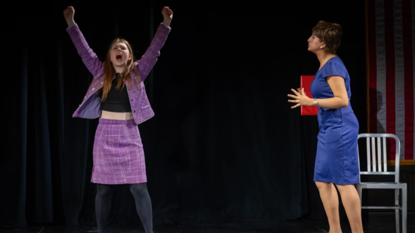 Two women on stage; one with arms raised and mouth open, wearing a purple checkered suit, and the other with hands near chest, wearing a blue dress, in front of a black curtain.