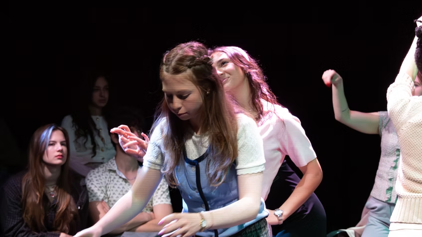 Multiple young women sitting and standing in a dark room, with one woman in the foreground reaching forward while others watch, some with serious expressions and one with a smiling face.