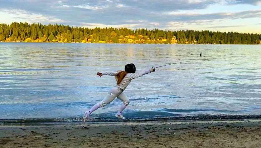 Person wearing a helmet and athletic clothing, holding a fencing blade, running along the beach near a lake with forested shoreline in the background.