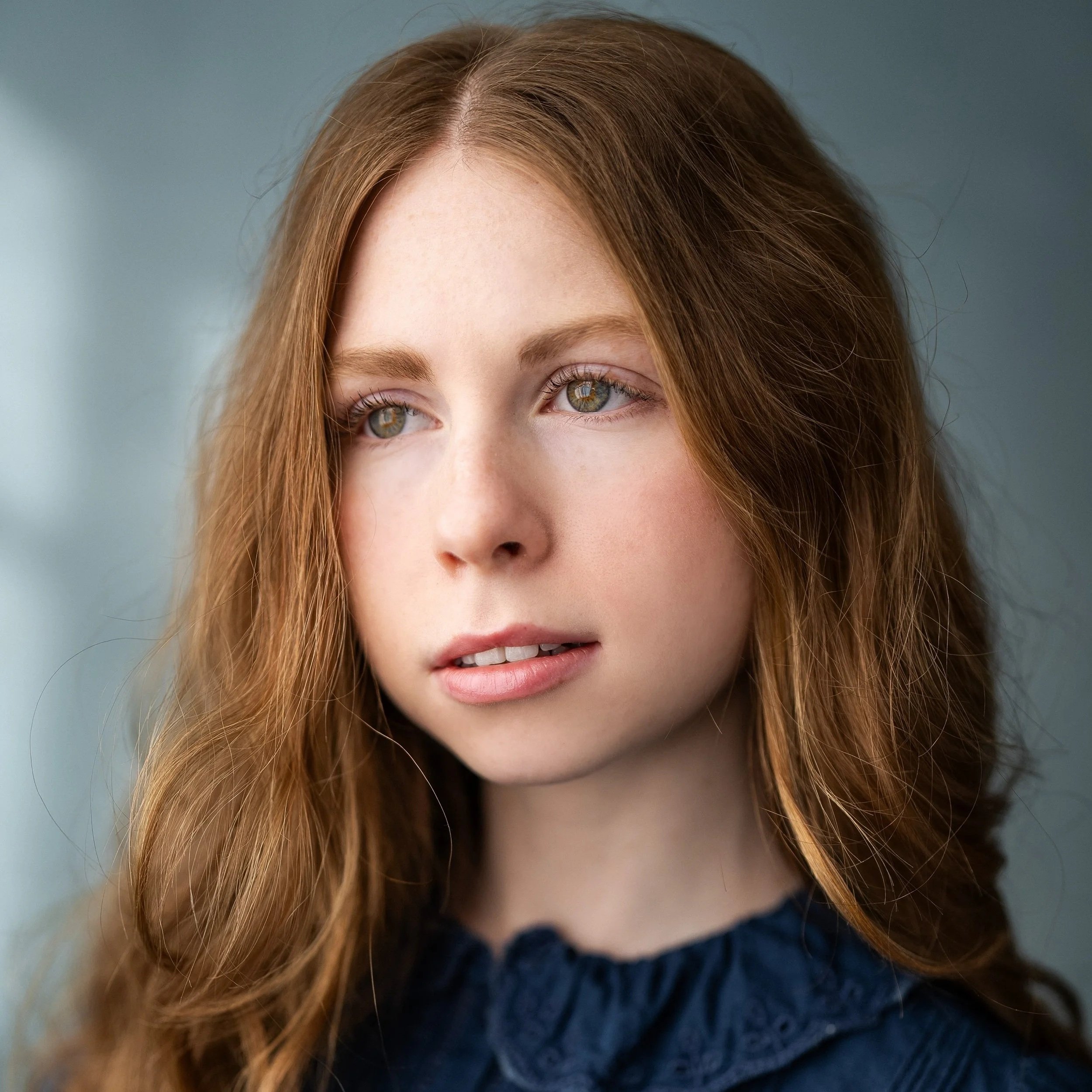 Close-up of a young woman with long, wavy red hair, fair skin, and blue eyes, looking slightly to the side with a neutral expression.