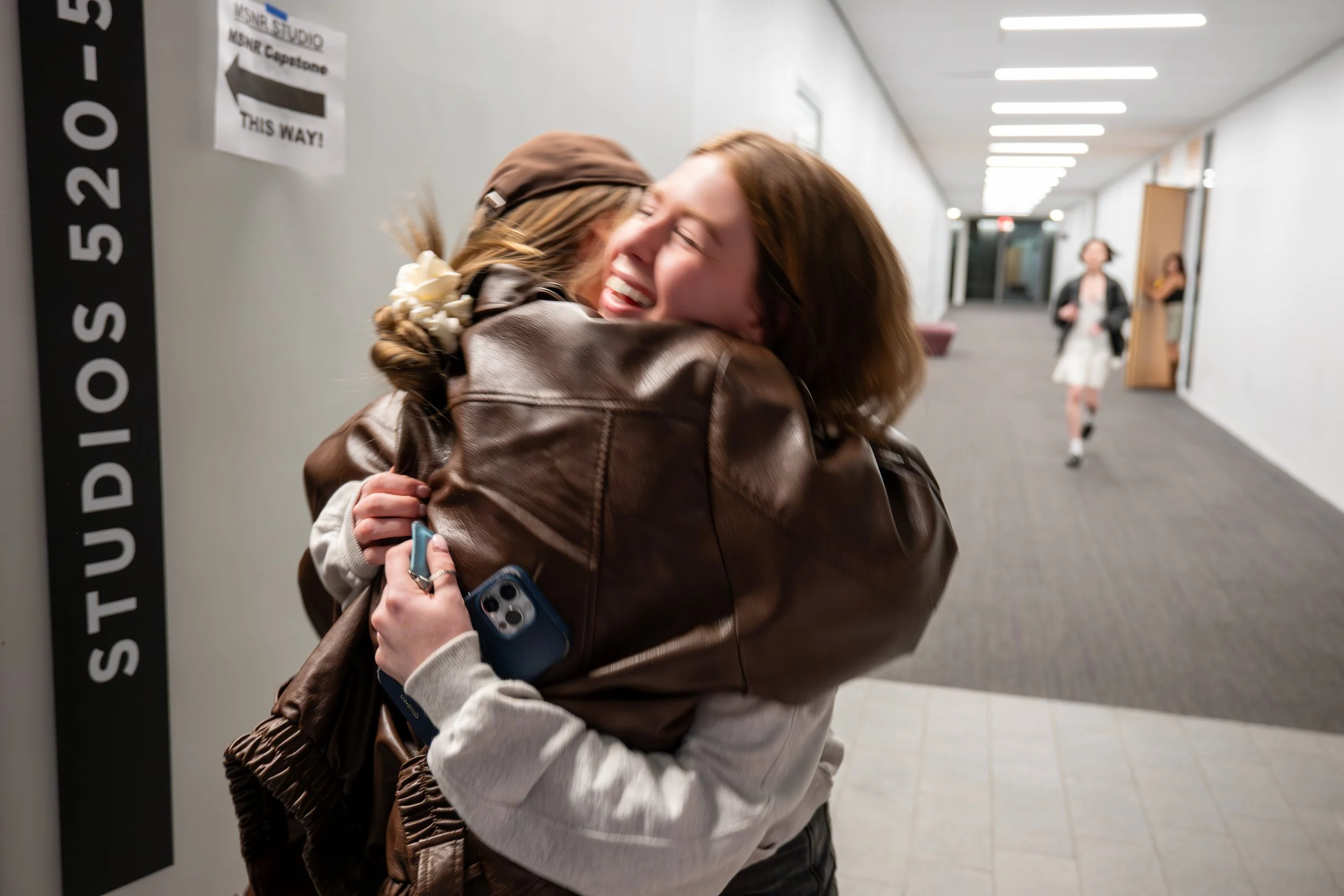 Two women hugging and smiling warmly in a hallway of a building, with a third woman walking in the background.
