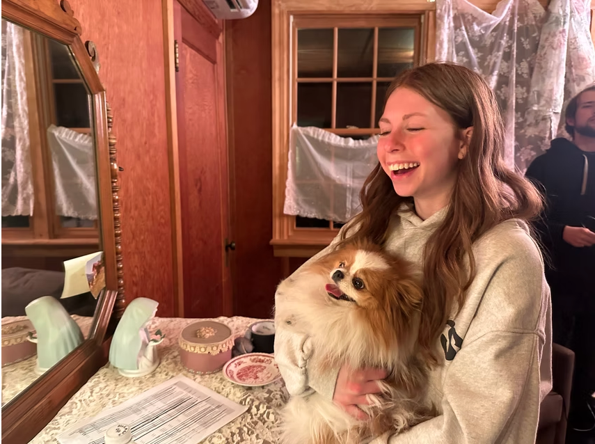 A young woman with long brown hair, smiling and laughing, is holding a small fluffy dog with a cheerful expression. The scene is inside a cozy, wood-paneled room with a dresser, a mirror, and windows with lace curtains.