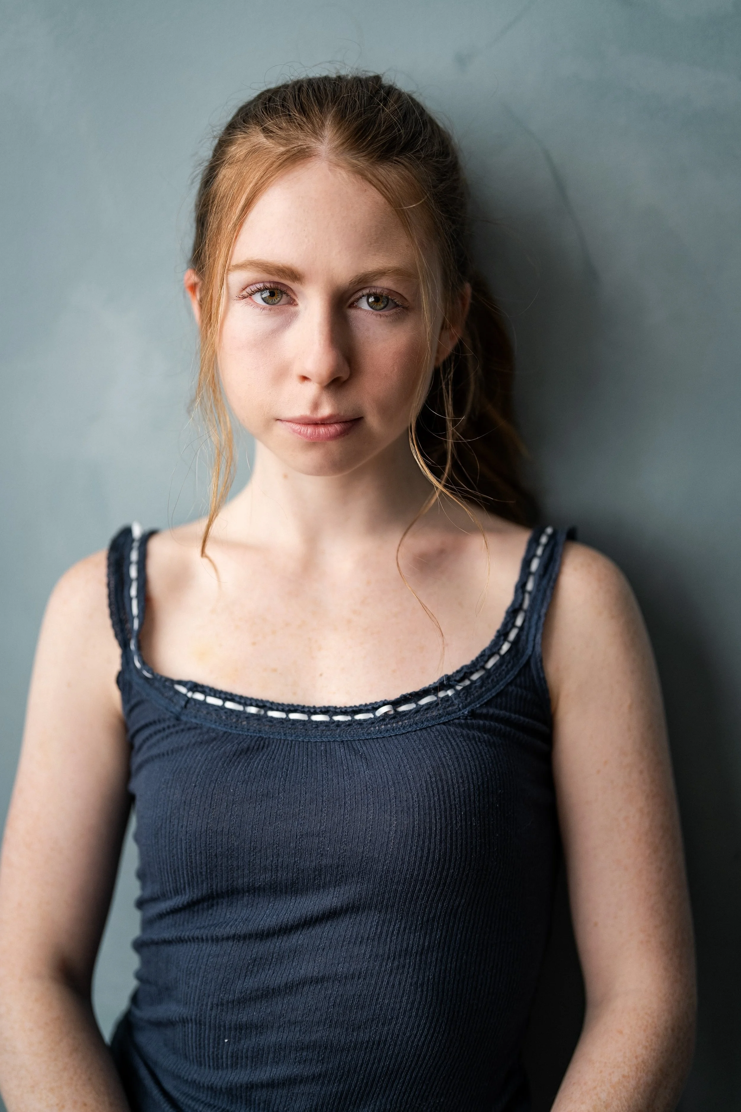 A young woman with red hair tied back, wearing a dark sleeveless top, standing against a plain wall. Pretty girl. 