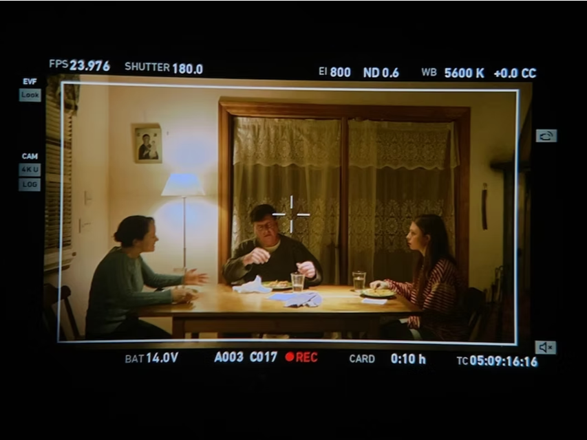Three people sitting at a dining table having a conversation, with a man in the middle, a woman on his right, and another woman on his left, in a warmly lit room with a wooden window and lace curtains.