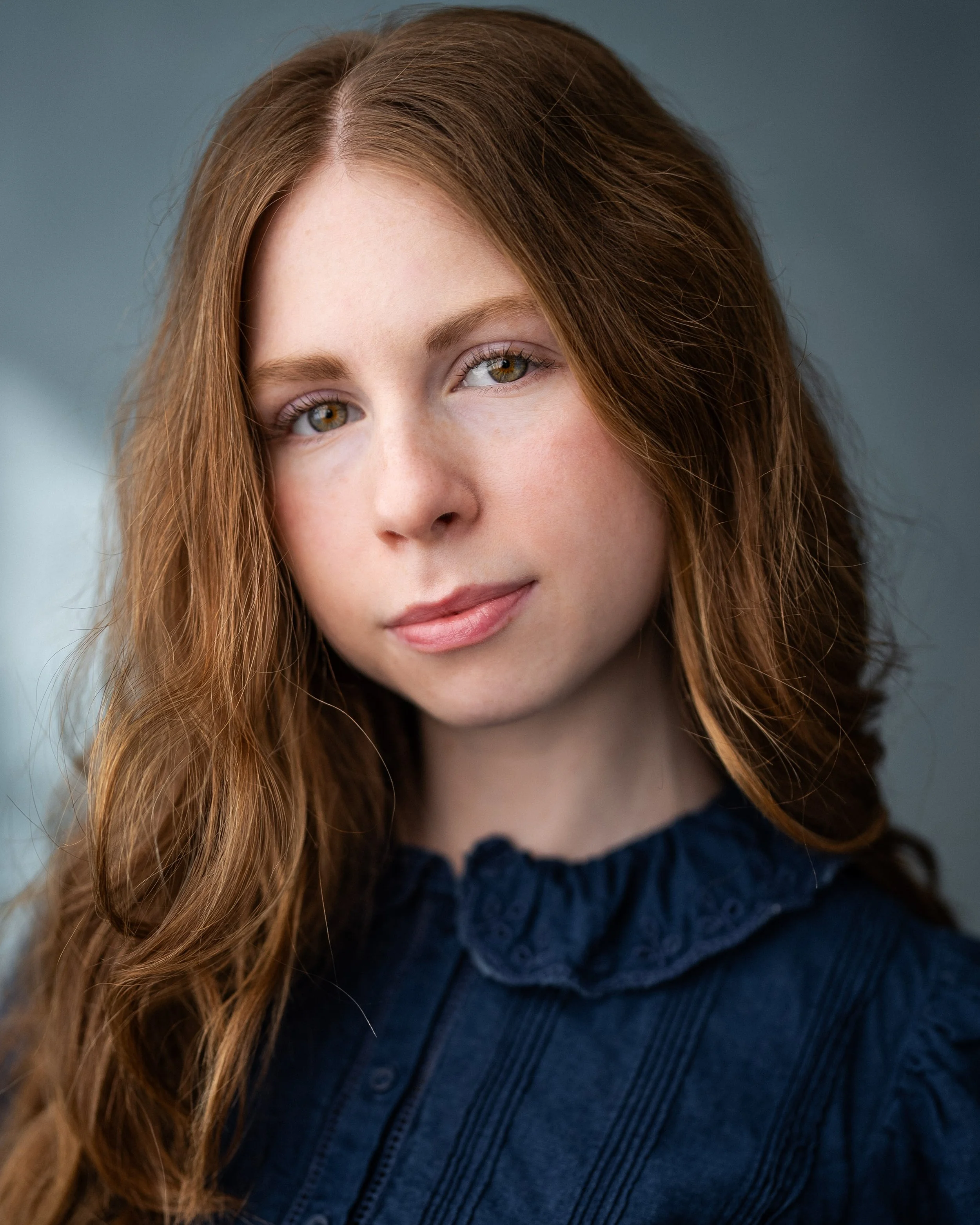 Close-up portrait of a young woman with long wavy red hair, fair skin, green eyes, and wearing a navy blue ruffled top. Actor. Merida from Brave.