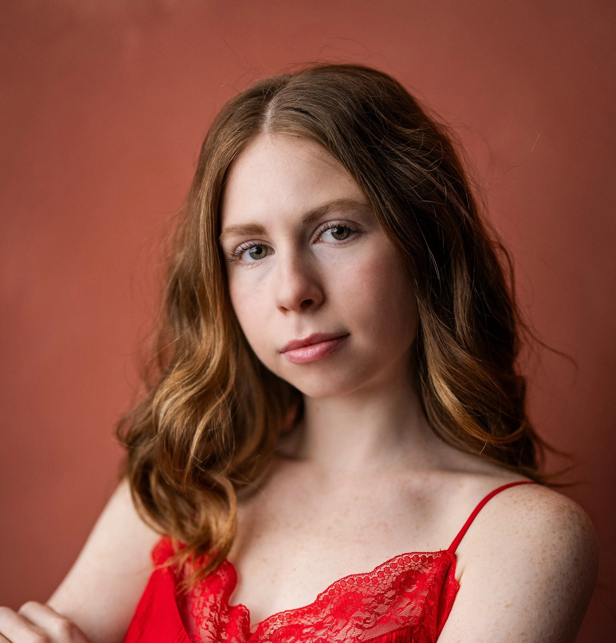 A young woman with wavy, shoulder-length auburn hair and light-colored eyes, wearing a red lace camisole, looking directly at the camera with a neutral expression, against a reddish-brown background.
