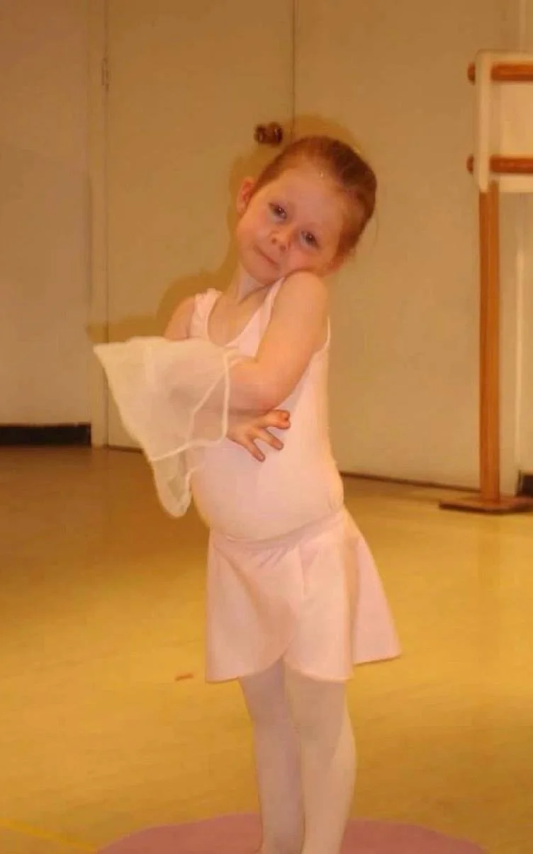 A young girl in a pink ballet outfit standing on a dance studio floor, with hands on her hips and a shy smile.