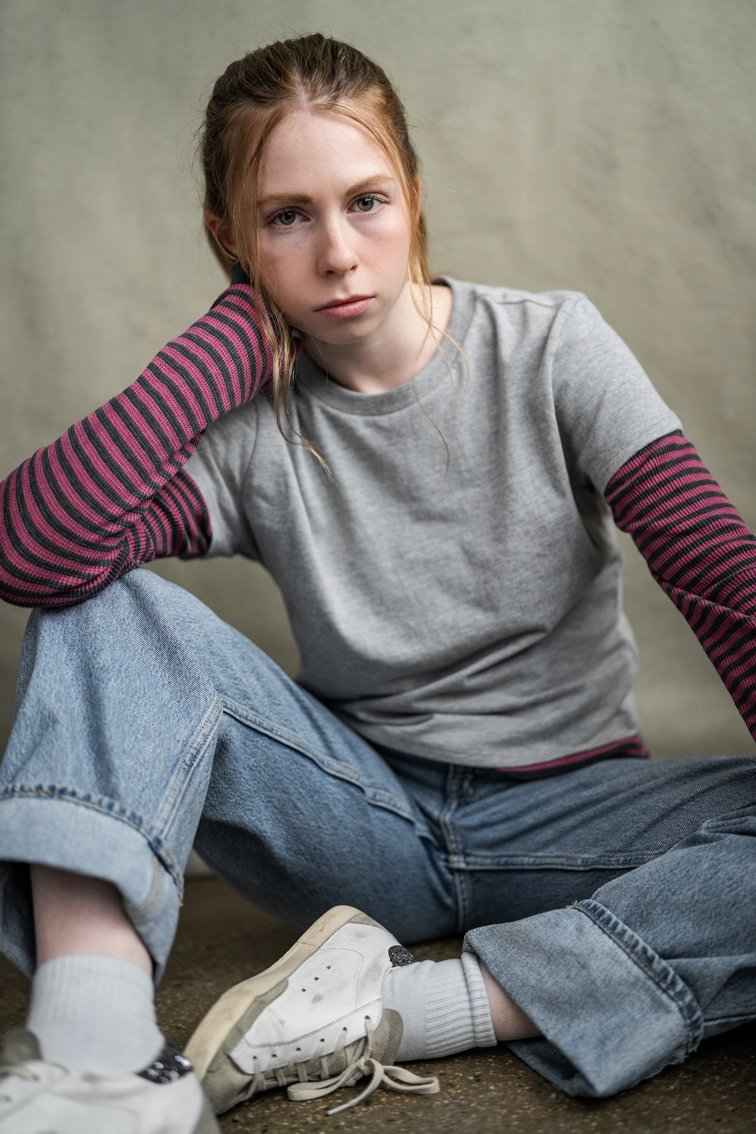 A young woman with red hair, wearing a gray t-shirt with striped pink and black sleeves, gray jeans, white sneakers, and white socks, sitting on the floor with a serious or contemplative expression.