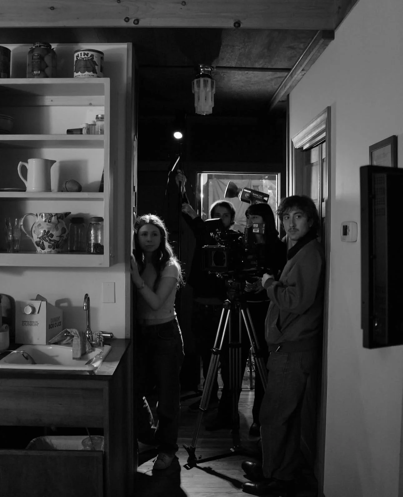A black and white photo of a film crew inside a kitchen, with a woman and a man near a camera and lighting equipment.