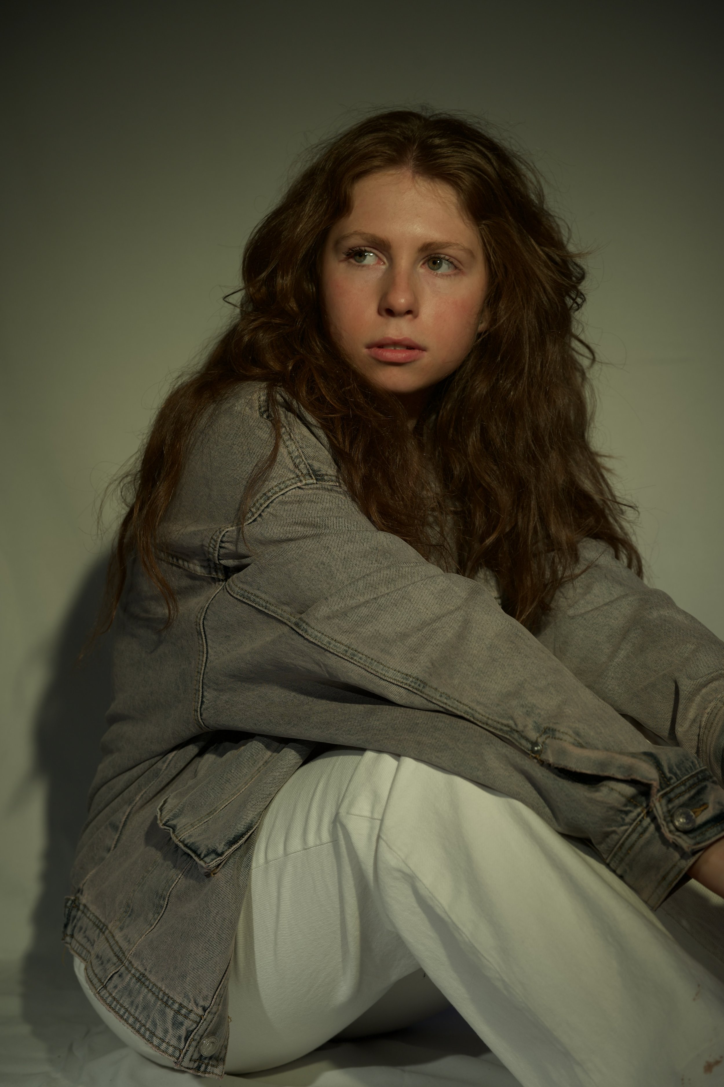 Portrait of a young woman with long, wavy light brown hair, wearing a gray denim jacket and off-white pants, sitting against a plain background. Merida from Brave.