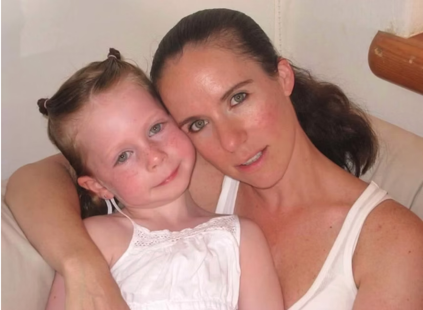A woman and a young girl sitting closely together on a beige couch, smiling and looking at the camera. The woman has dark hair pulled back, and the girl has curly red hair tied in pigtails. Both are wearing white tops.