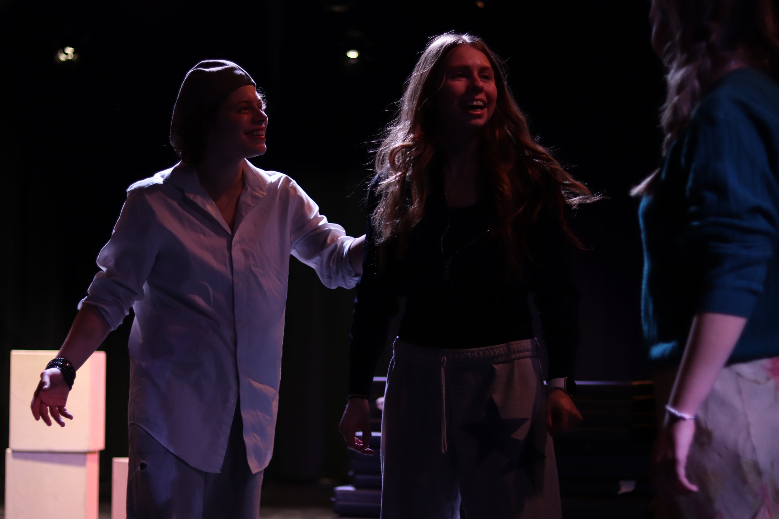 Three women are having a conversation on a dark stage, with one woman placing her hand on another woman's shoulder, all smiling.
