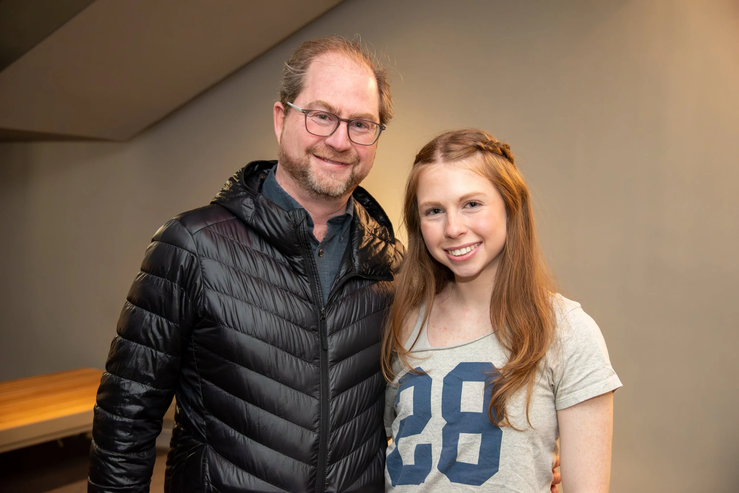 A man with glasses and a beard wearing a black puffer jacket, standing next to a young woman with long red hair in a grey T-shirt, both smiling at the camera indoors.