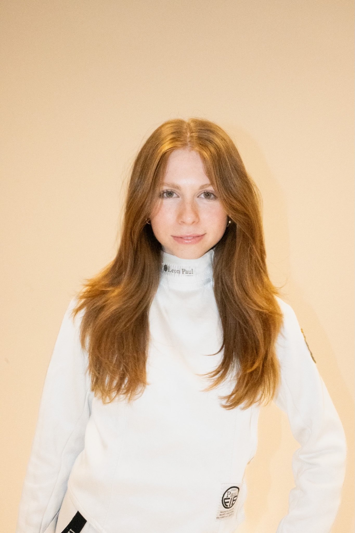 A young woman with long red hair wearing a white fencing uniform with a high collar and logo patches, standing against a plain beige background.