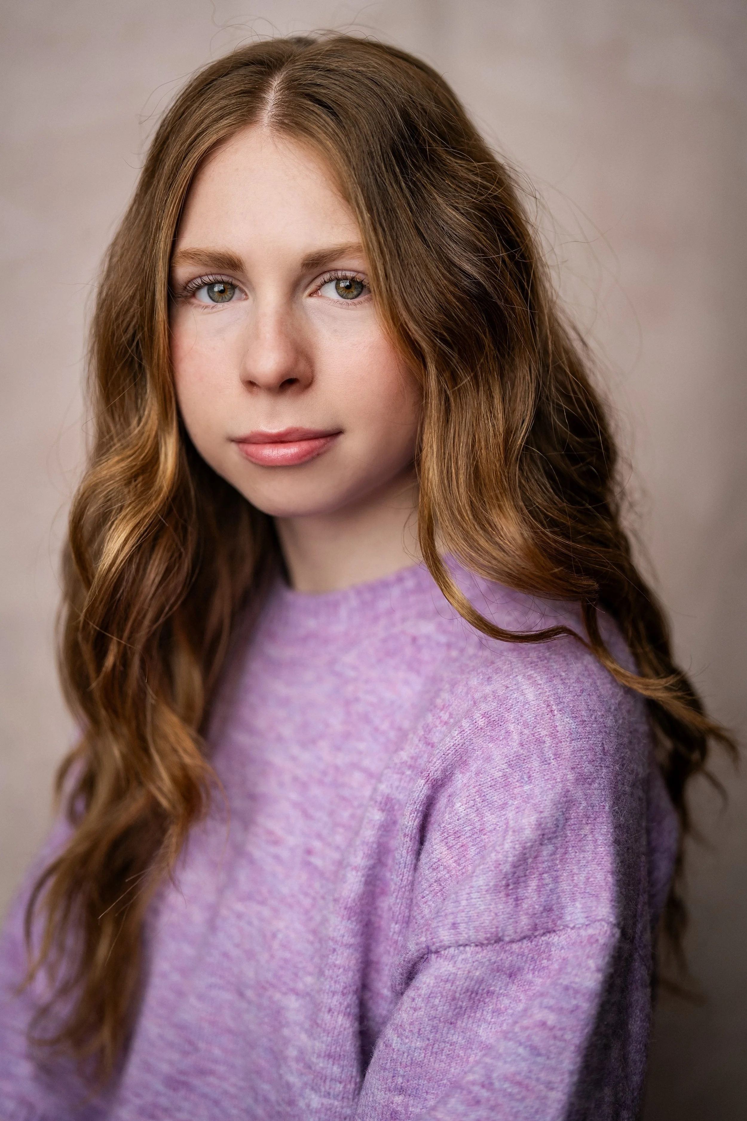 A young woman with long, wavy red hair and light-colored eyes, wearing a purple sweater, looking at the camera with a neutral expression against a plain beige background.