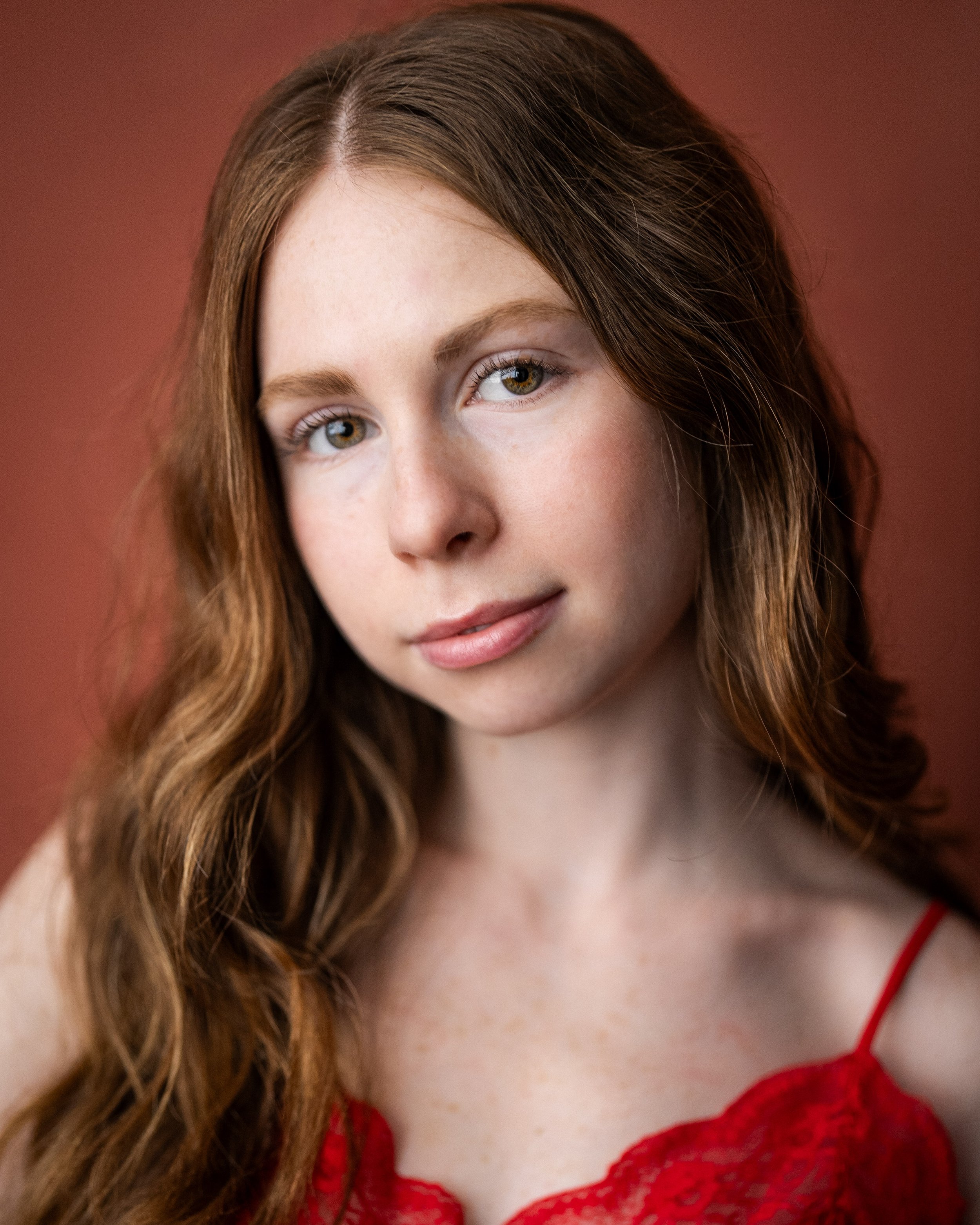Close-up portrait of a young woman with long wavy red hair, fair skin, and light-colored eyes, wearing a red lace top, against a reddish background.