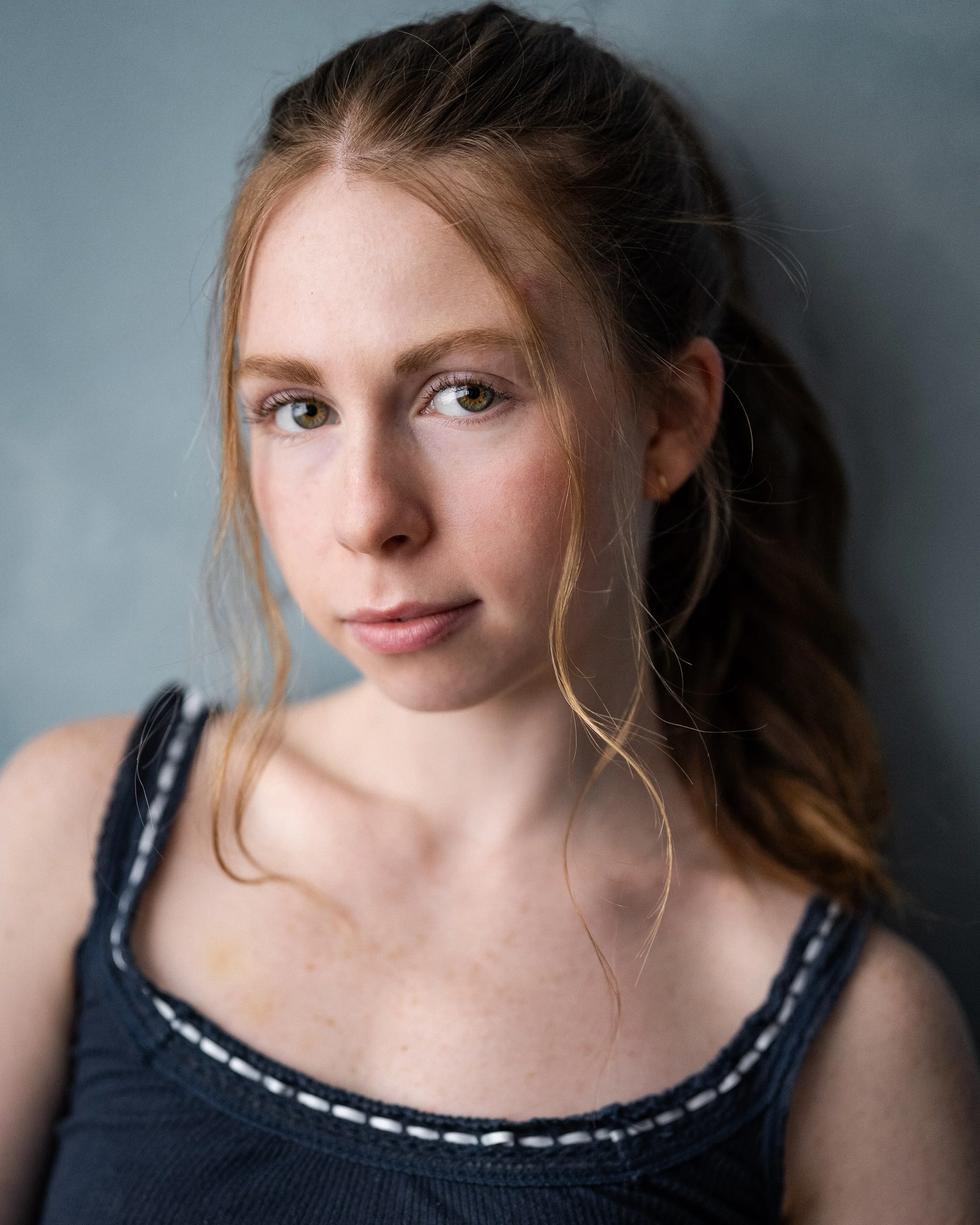 A close-up portrait of a young woman with red hair tied back, wearing a navy tank top, standing against a grey wall. Pretty girl.