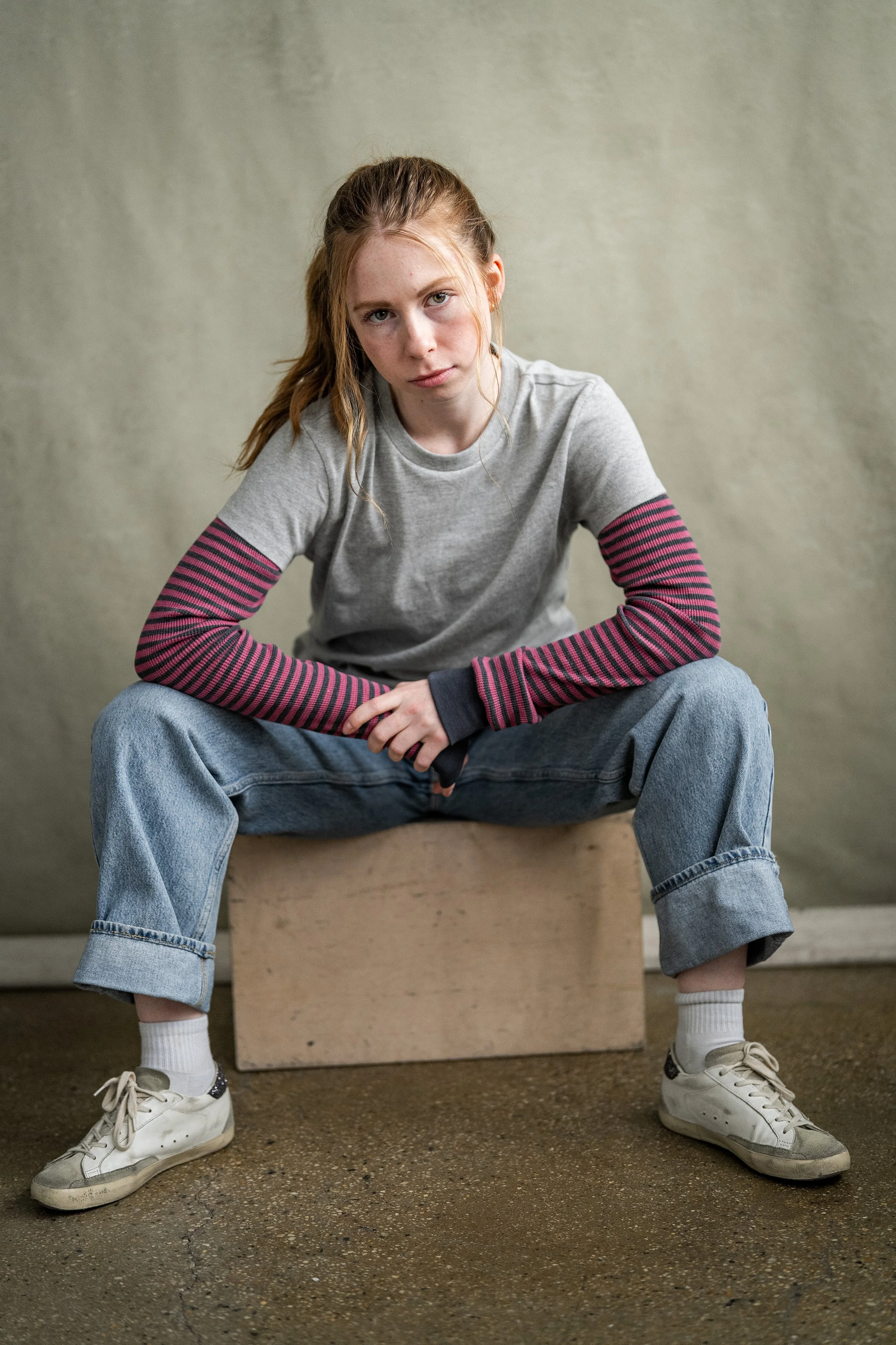 A teenage girl with reddish hair sitting on a wooden box in front of a plain wall, wearing a gray t-shirt with striped long sleeves underneath, loose jeans, white socks, and sneakers. Stranger Things. Teenage Actor.
