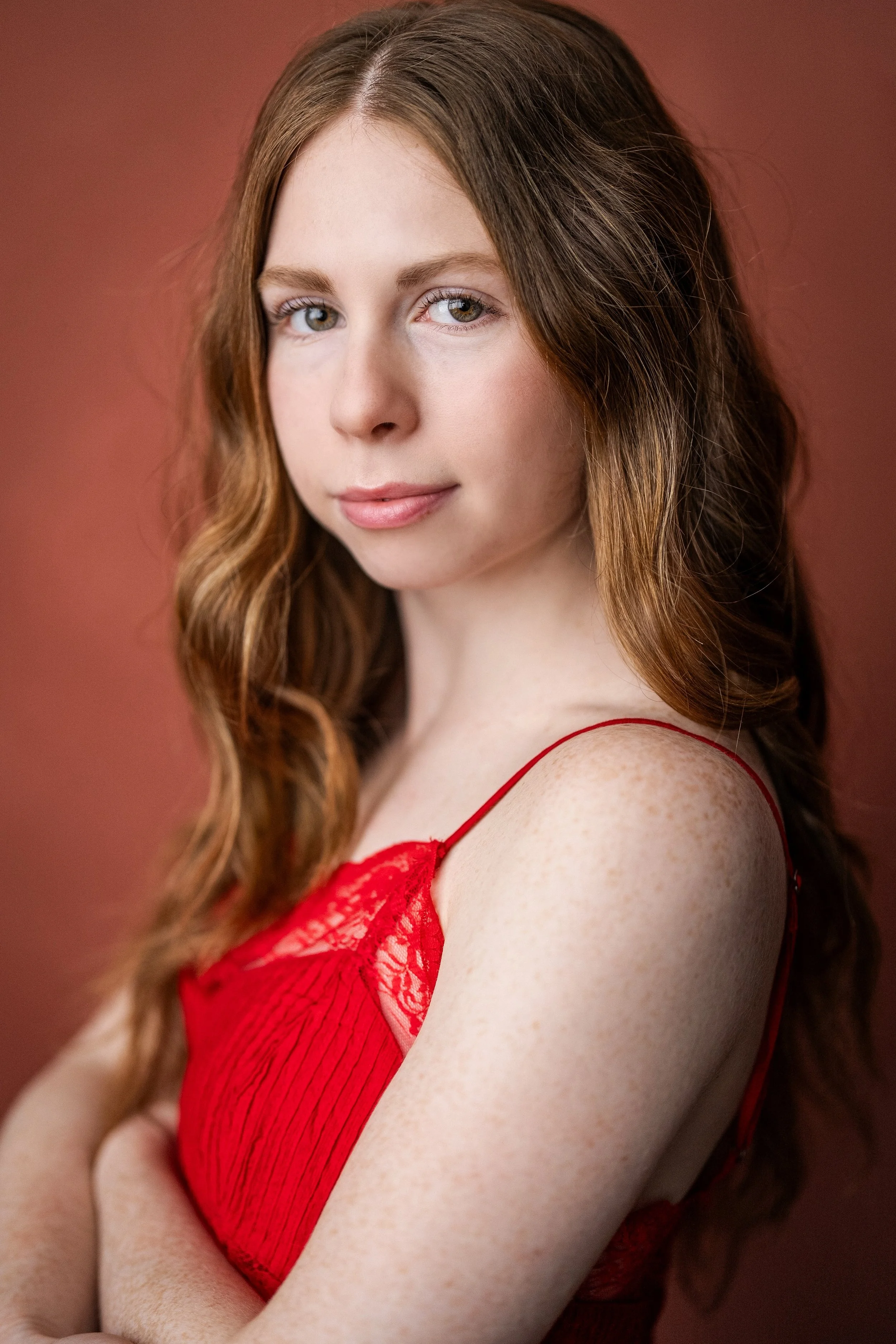 Portrait of a young woman with long wavy auburn hair, light skin with freckles, wearing a red lace spaghetti strap top, looking at the camera with a subtle smile against a solid red background. Pretty girl. 