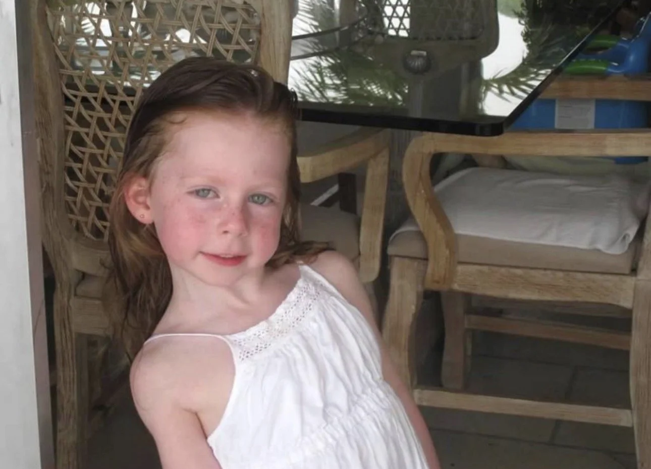 A young girl with red hair and freckles, wearing a white sleeveless dress, sitting outdoors on a wicker and wooden chair, with a glass table and chairs in the background.