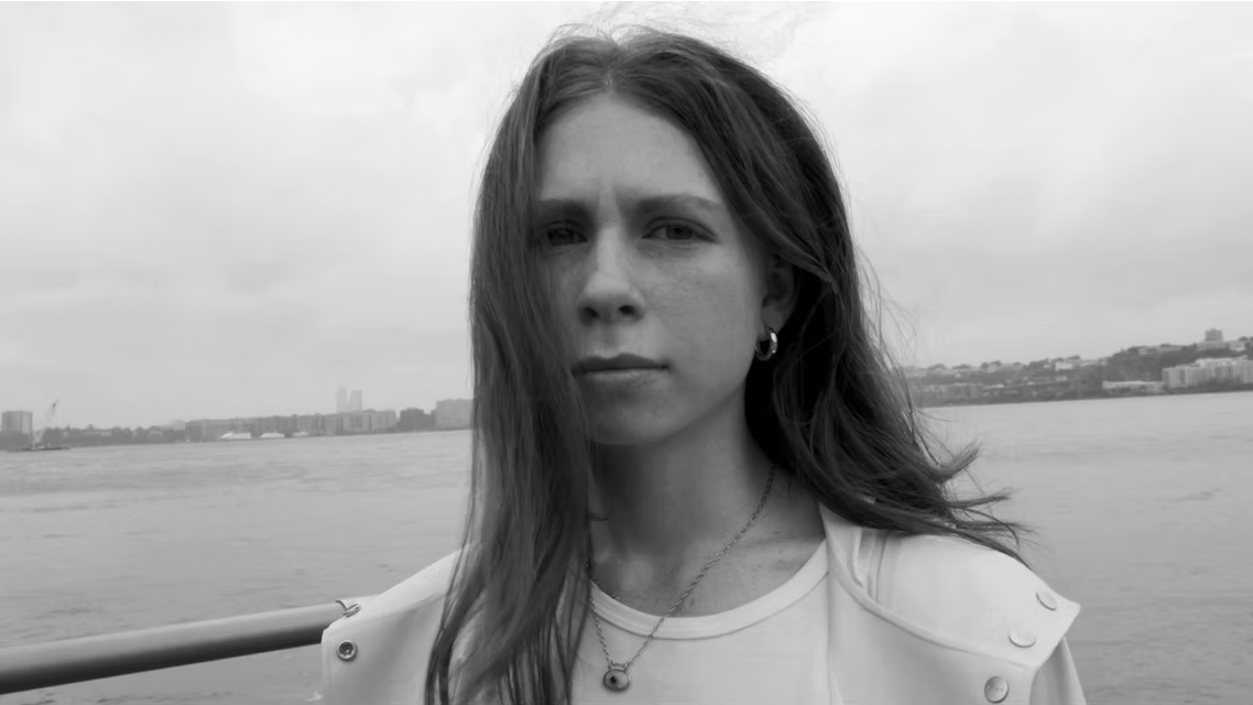 Black and white photo of a woman with long hair standing outdoors near a body of water, with a cityscape in the background.