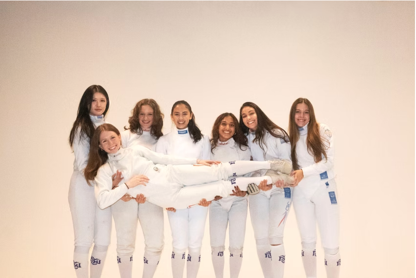 Group of seven women in white fencing uniforms, all smiling, holding up a woman in fencing gear horizontally in front of a plain beige background.