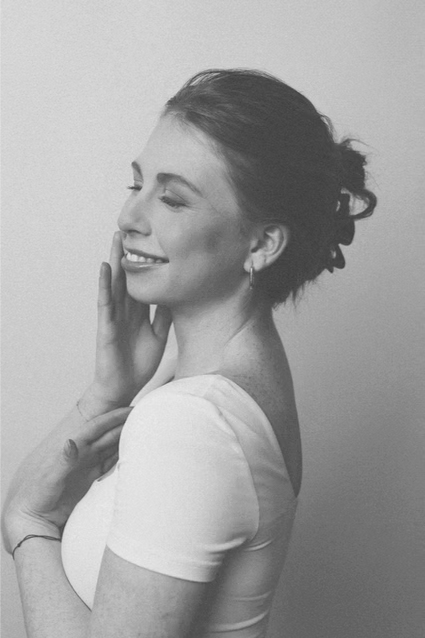 A black and white photo of a woman with her eyes closed, smiling gently, touching her face with one hand, and wearing a short-sleeved top and small earrings.