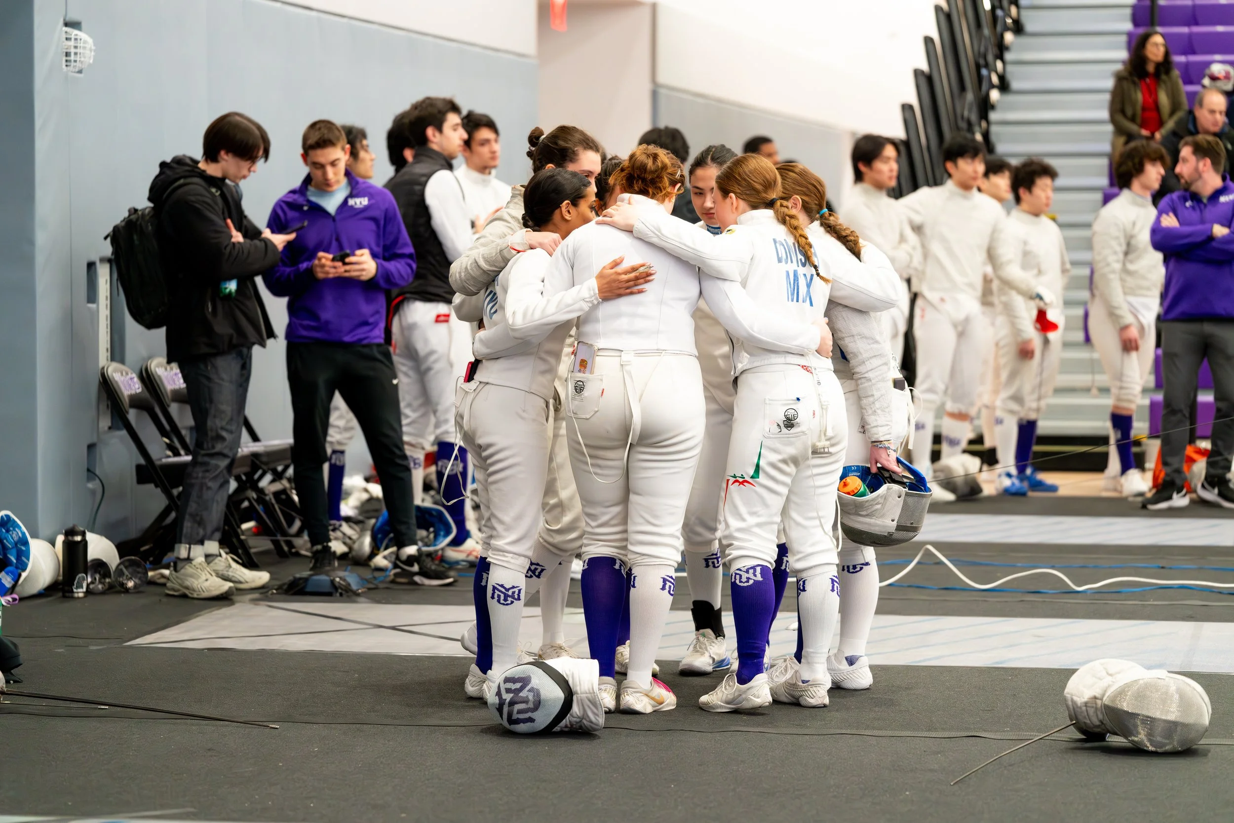 College women's fencing team huddling together in a gym area, with fencing gear and equipment around them, before a match.