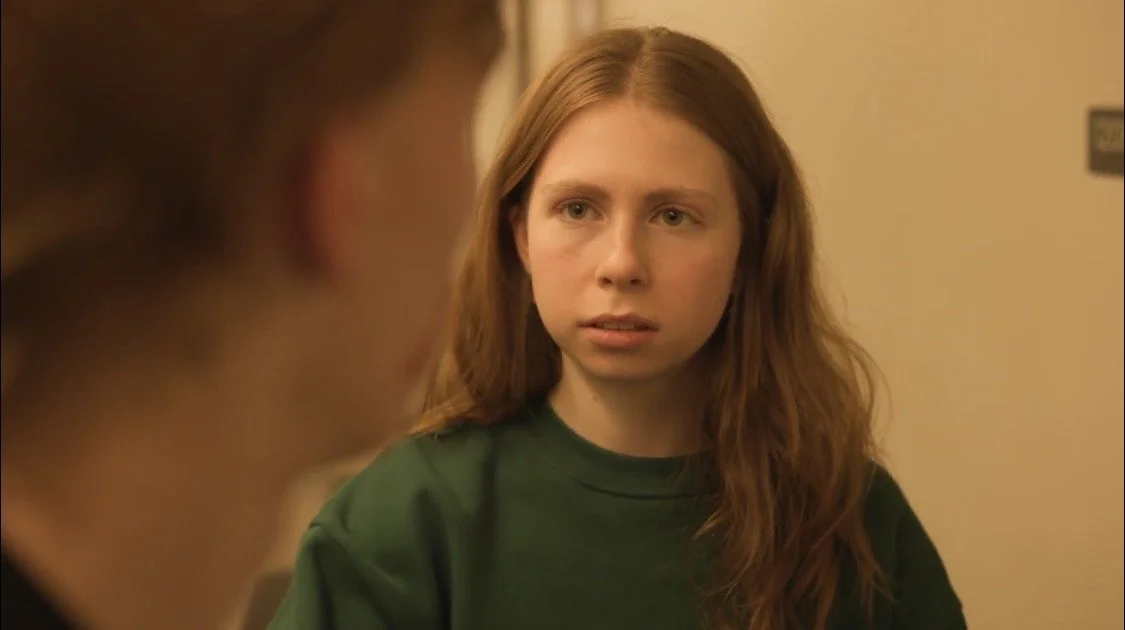 A young woman with long red hair wearing a dark green shirt, looking at someone off-camera with a serious expression.