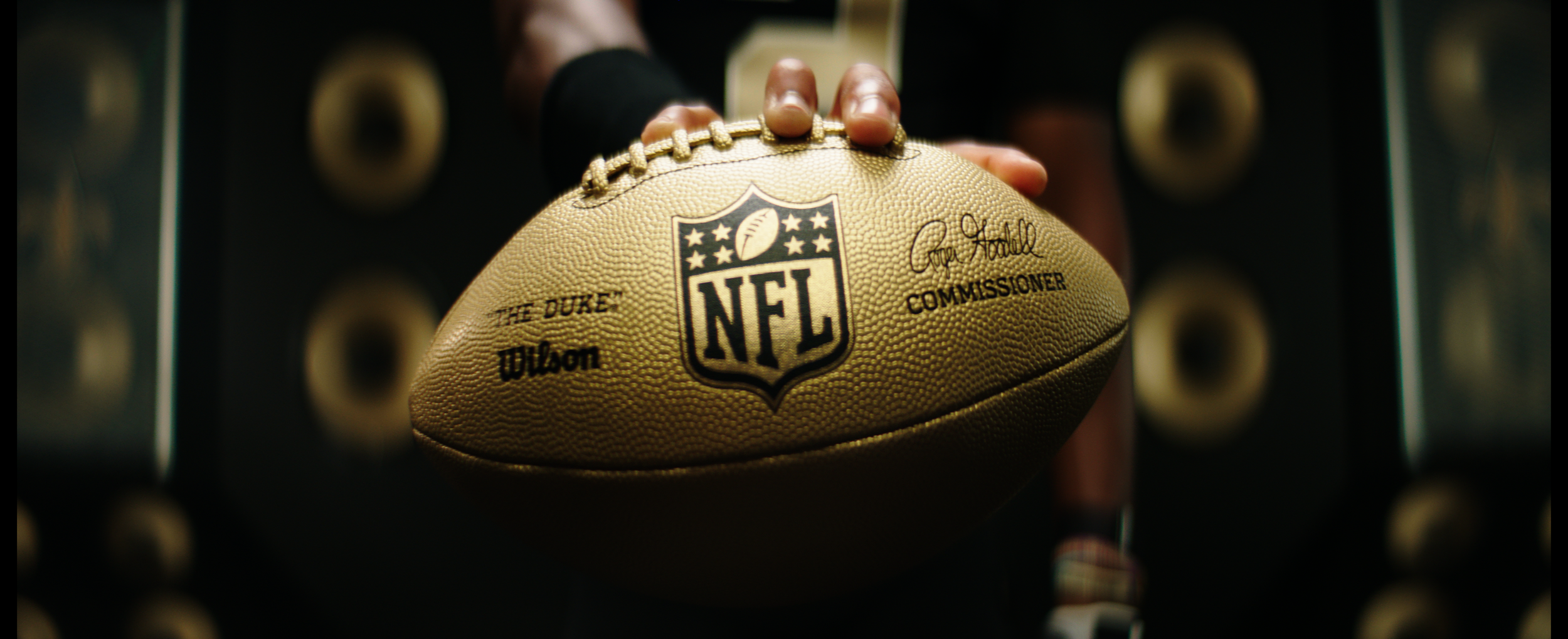 Close-up of an NFL gold football with logos and signatures. New Orleans Saints player wearing a helmet and jersey number 13 stands in front of a wall filled with large gold speakers. New Orleans Saints. Shot on Alexa Mini with anamorphic lenses.