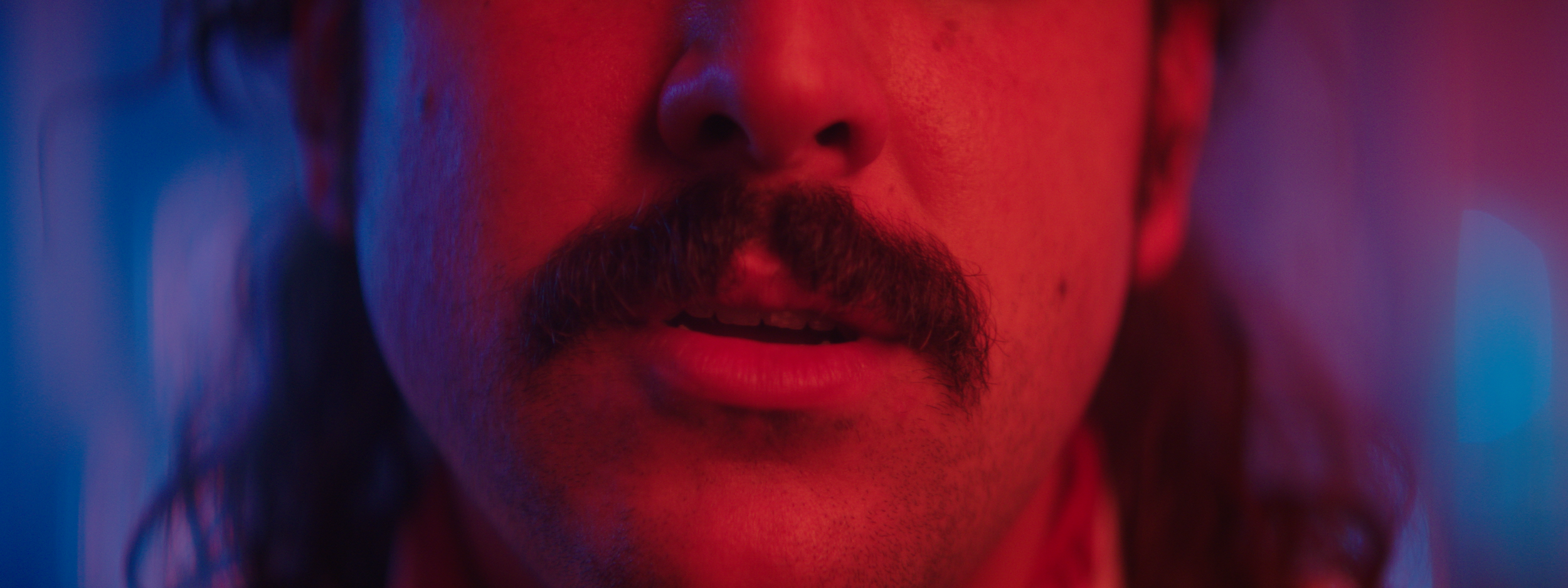 Close-up of a person's lower face with a mustache, illuminated by blue and red lighting.