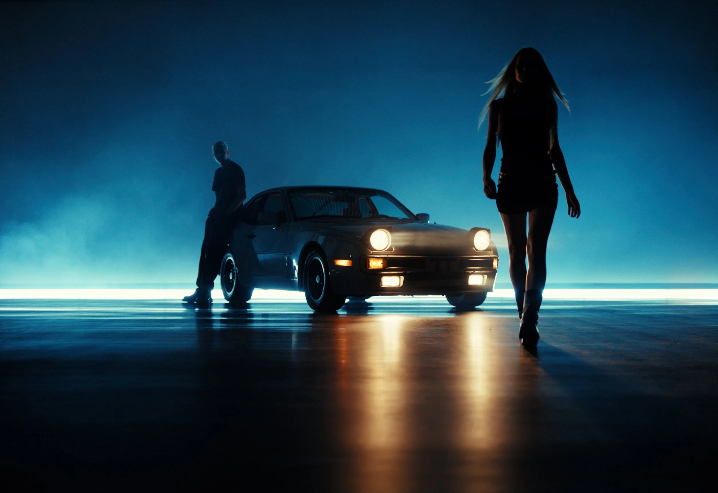 A woman walking in front of a classic sports car with the headlights on, with a man leaning against the car in the background, set against a dark, atmospheric background with glowing lights.