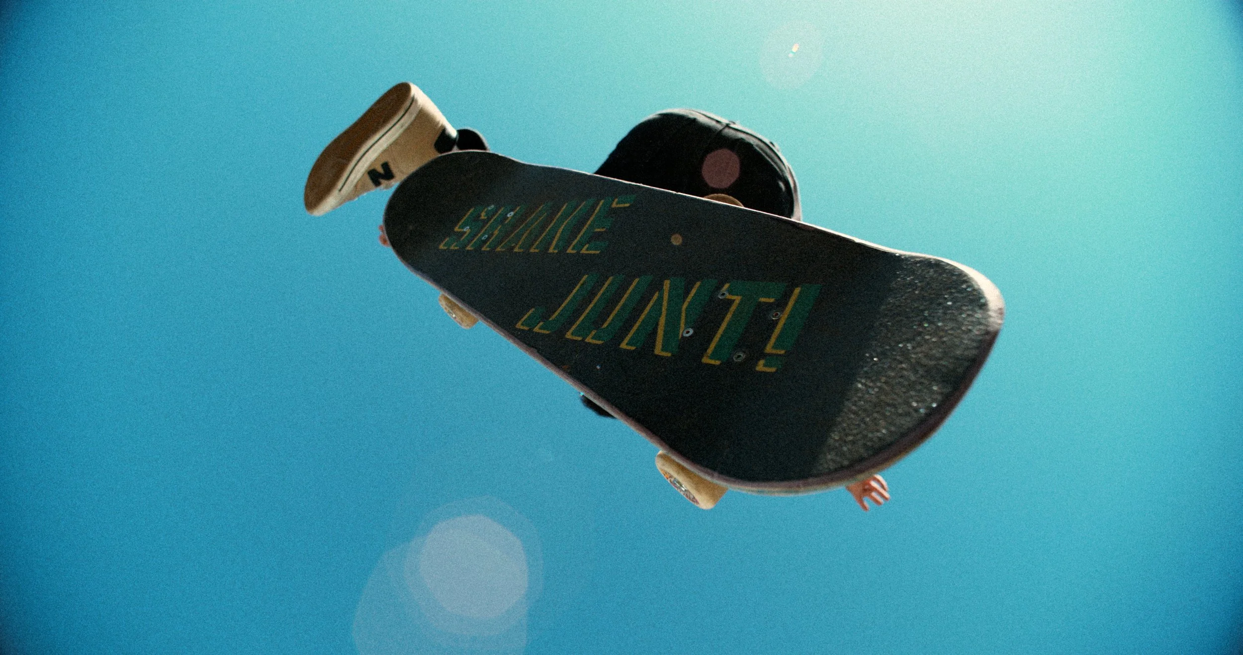 Low angle view of a skateboarder performing a trick in the air, with "SHINE JUNT!" on the skateboard's deck against a clear blue sky.