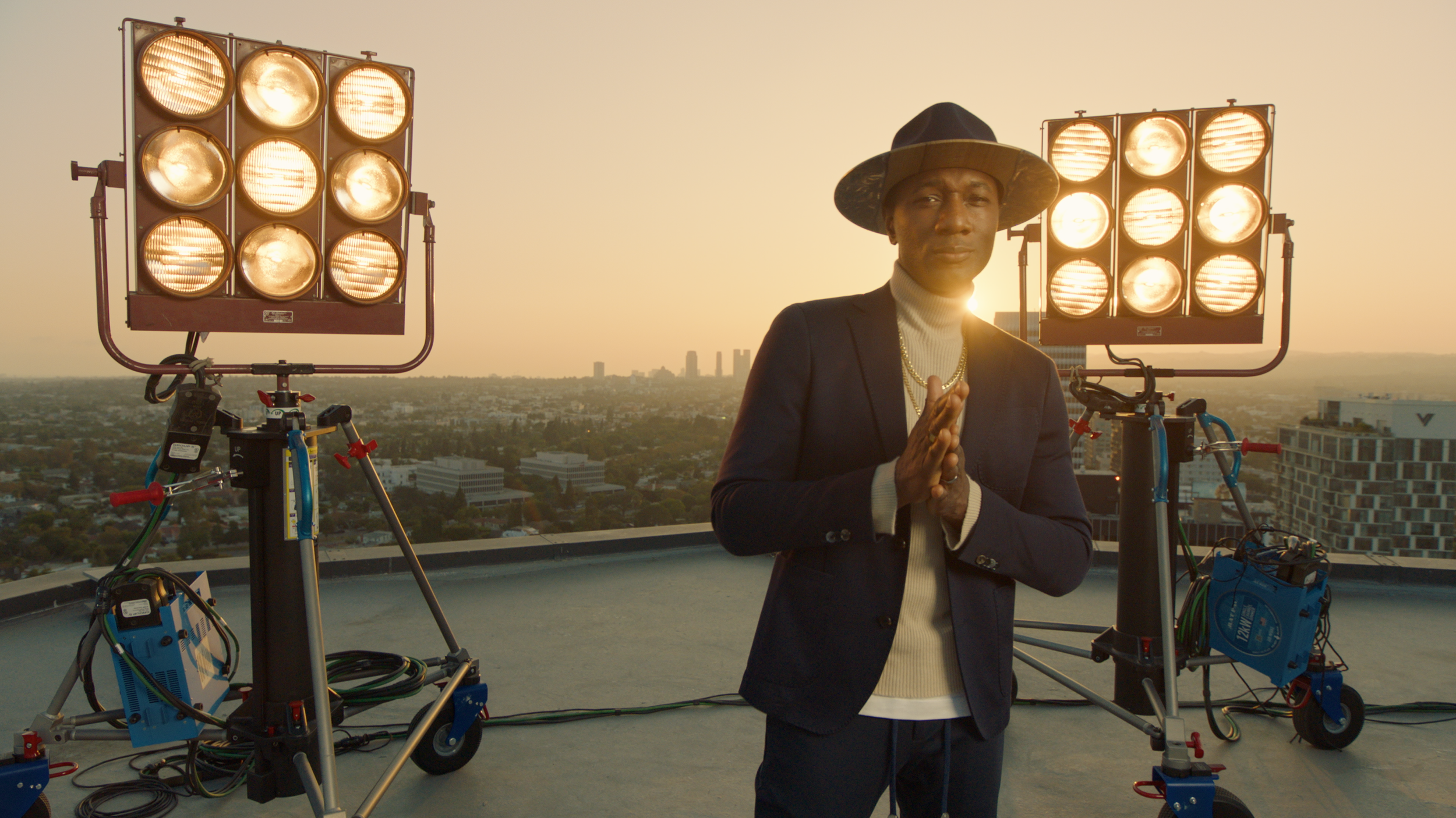 Man in hat and suit standing on rooftop with film lights during sunset