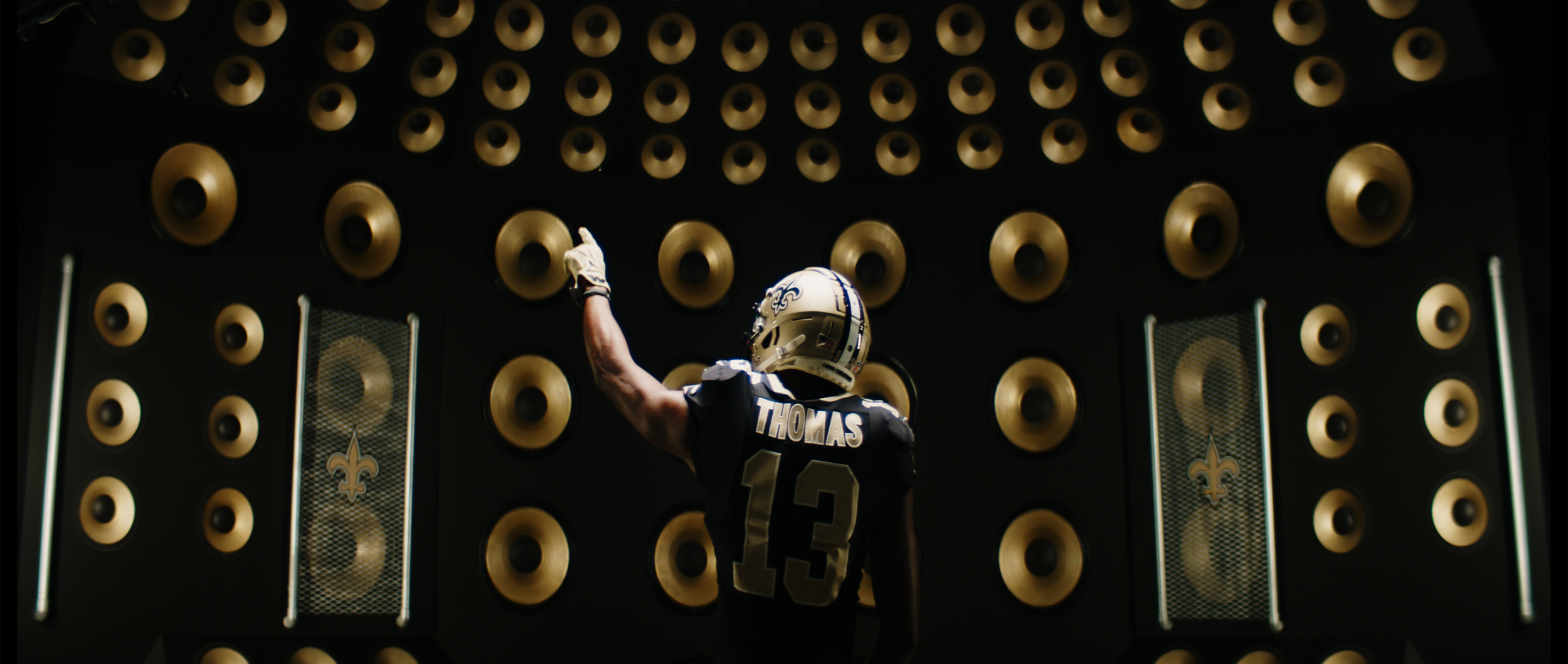 New Orleans Saints player wearing a helmet and jersey number 13 stands in front of a wall filled with large gold speakers. New Orleans Saints. Shot on Alexa Mini with anamorphic lenses.