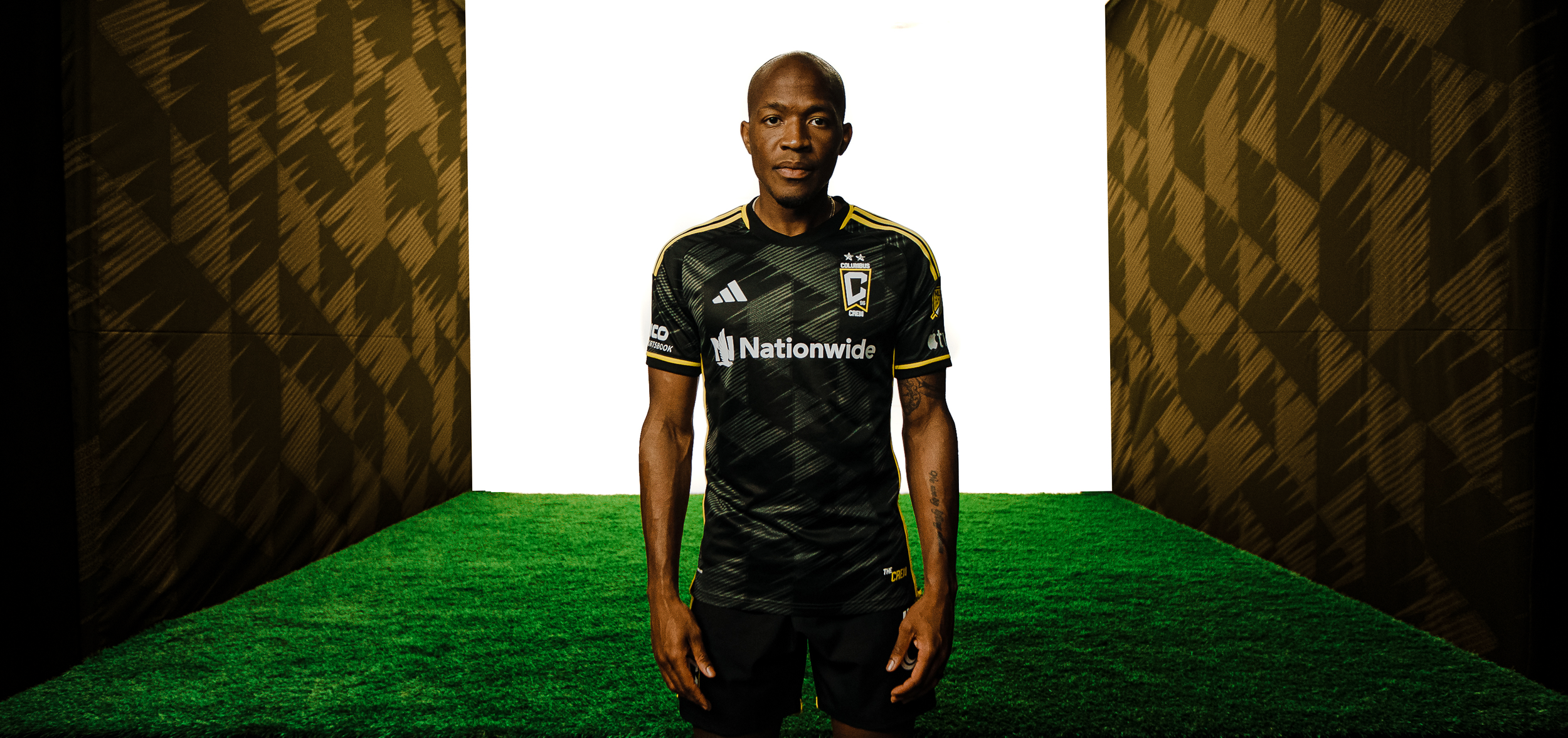 Soccer player wearing a black jersey with yellow accents and branding, standing on artificial grass with patterned walls.
