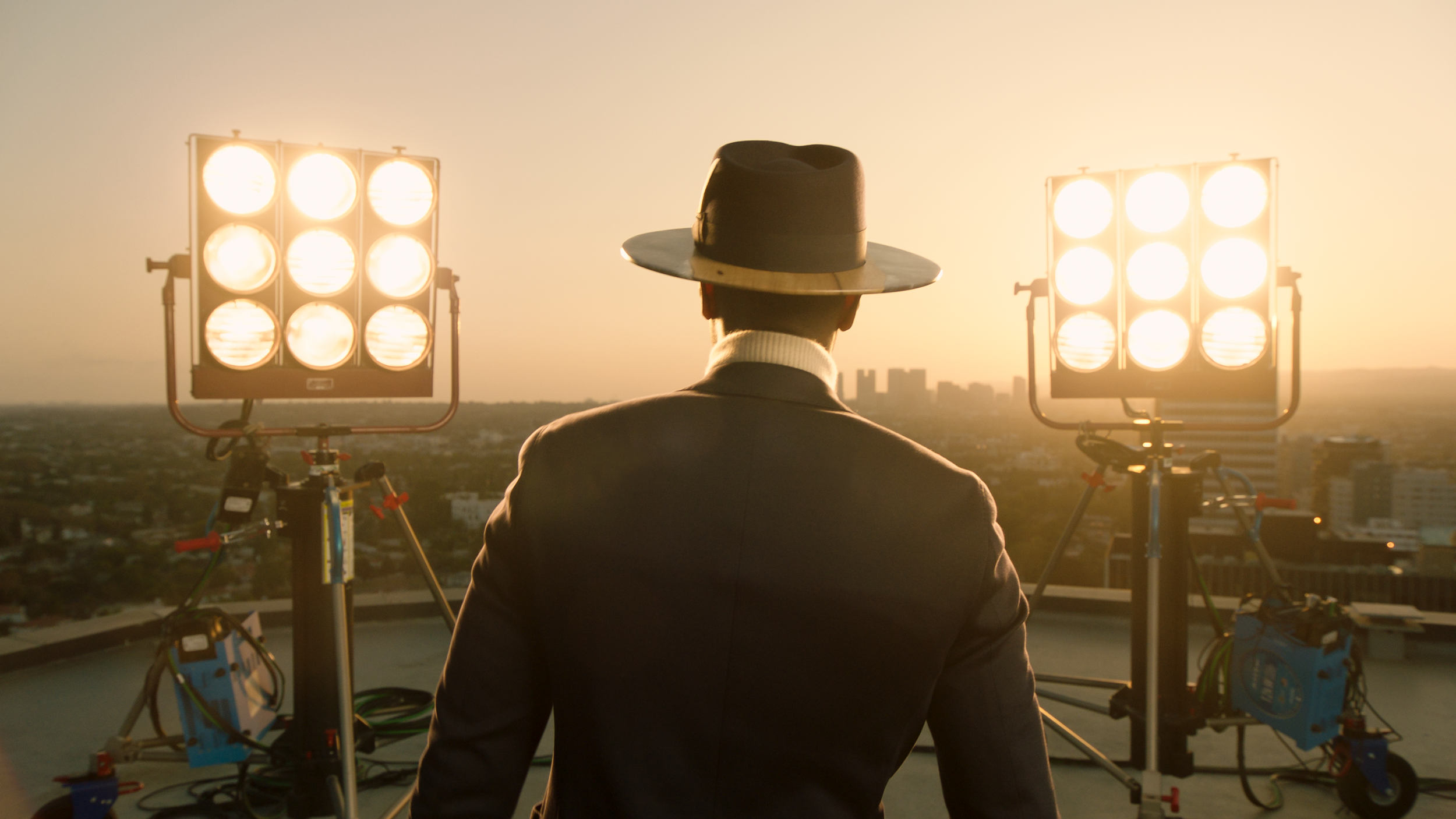 Person in a suit with a fedora stands on a rooftop with large studio lights at sunset, overlooking a cityscape.