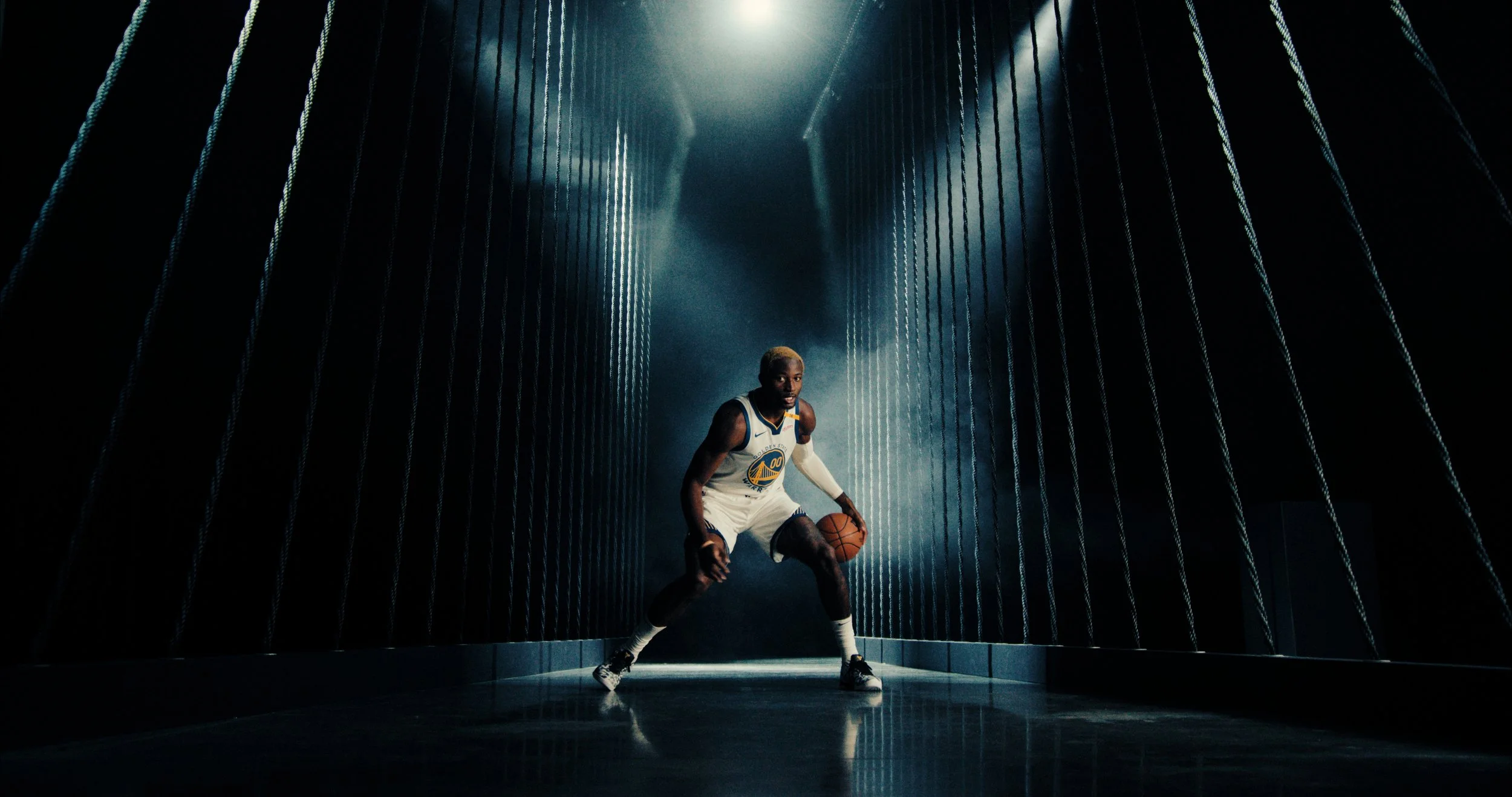 Basketball player in Golden State Warriors uniform dribbling a basketball in a dimly lit hallway.