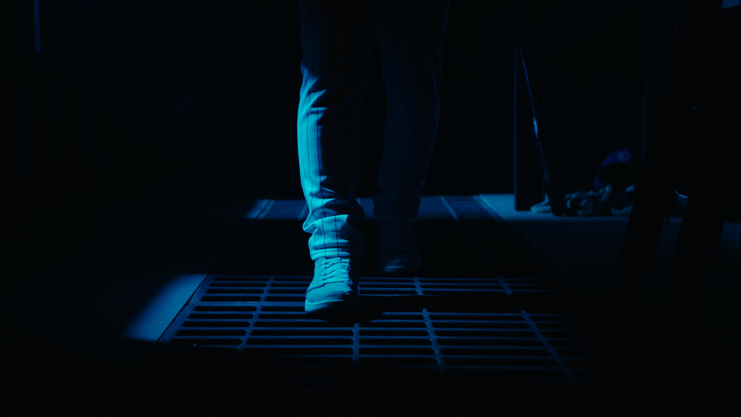 Person walking on a dark, grid-like floor wearing light-colored sneakers and striped pants, illuminated by blue lighting.