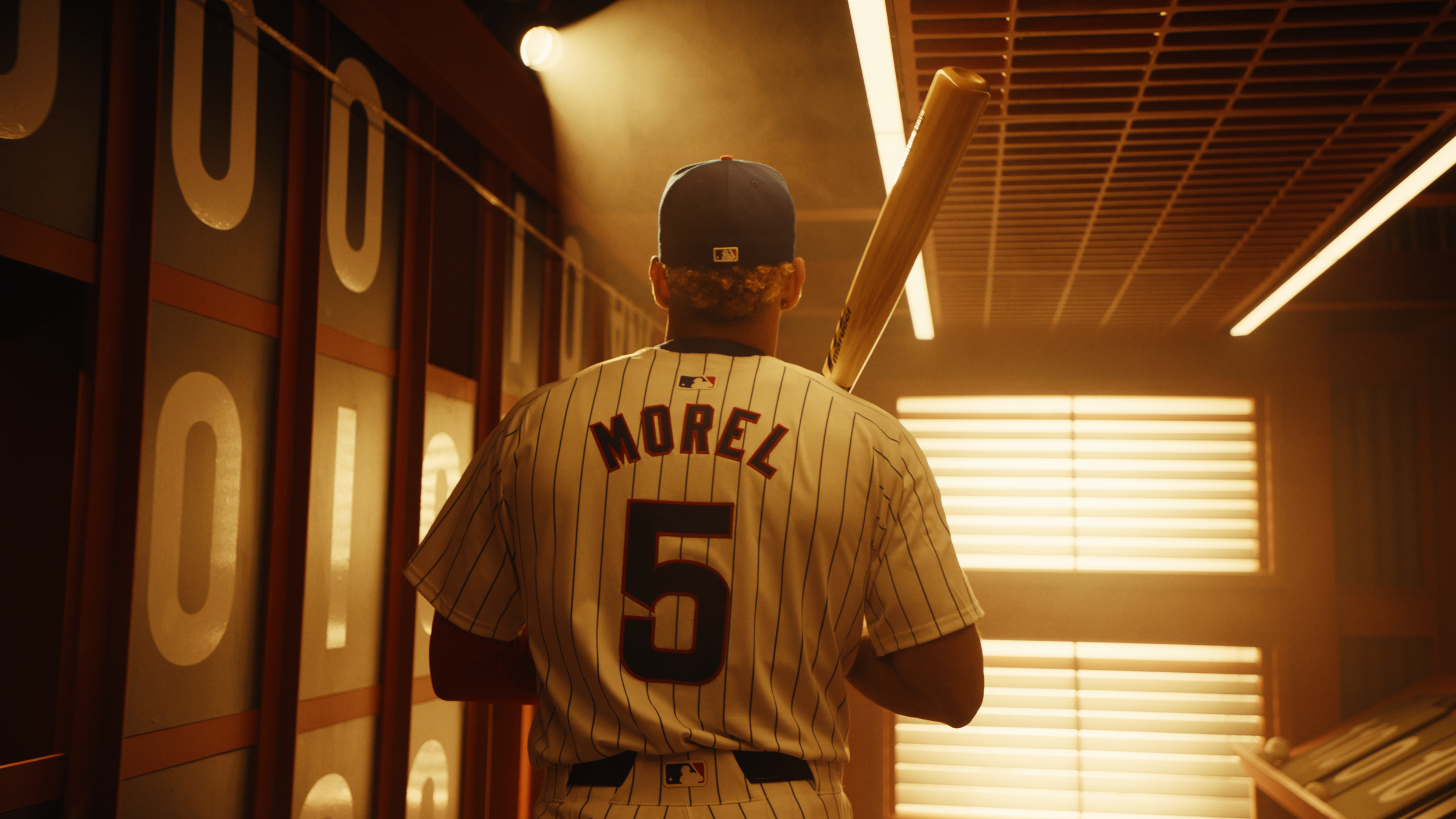 Back view of a baseball player wearing a pinstripe jersey with the name 'Morel' and number 5 on it, holding a bat over his right shoulder, standing in a room with warm lighting and window blinds.