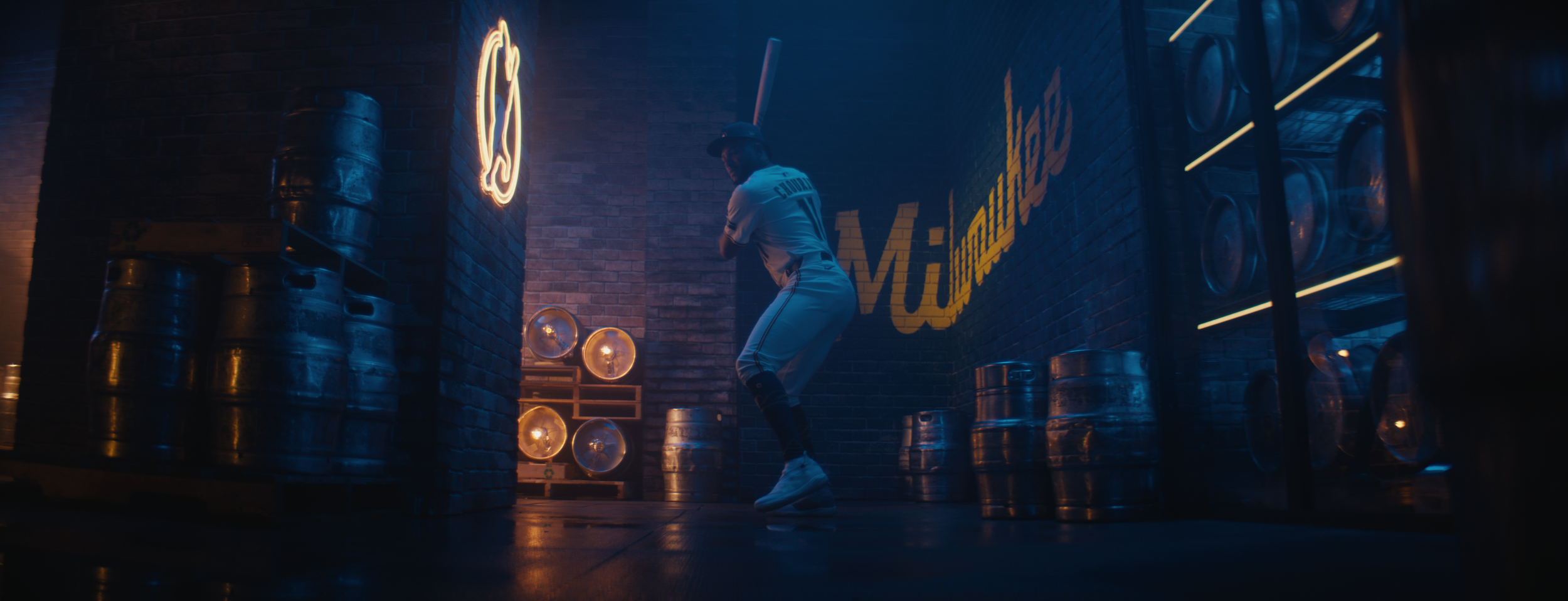 A person in a baseball uniform practicing with a bat in a dimly lit indoor sports facility, with beer kegs stacked and a neon sign on the wall.