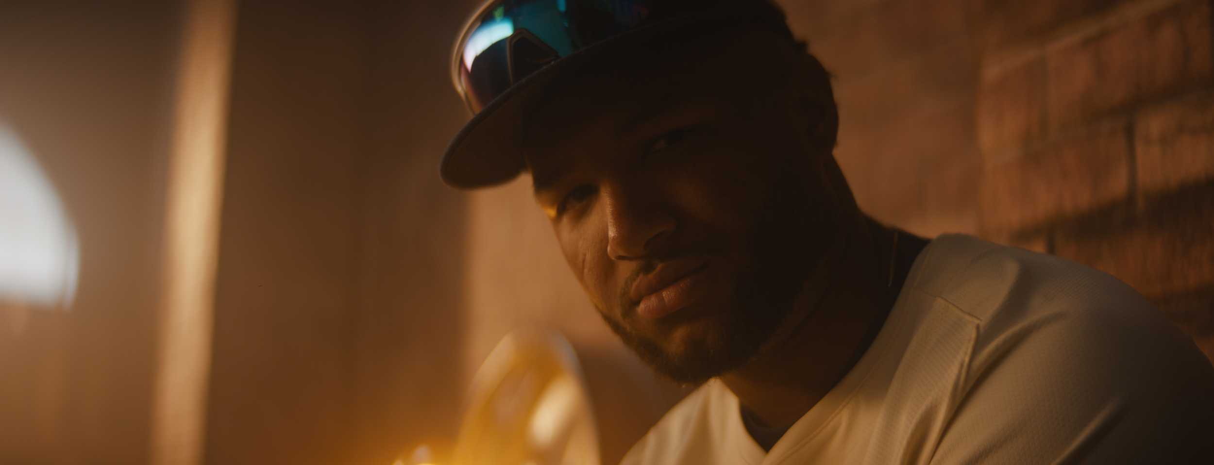 A man wearing a cap with sunglasses resting on it, looking into the camera with a slight smile, in a dimly lit room with a brick wall background.