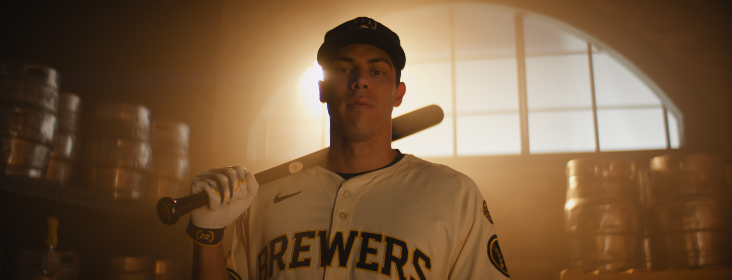 A baseball player holds a bat over his shoulder in a warehouse with stacked barrels, wearing a Brewers jersey and cap, backlit by sunlight.